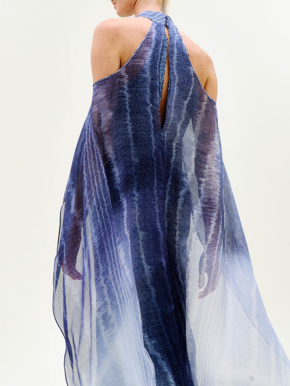 A person is seen from behind in the Serenela Dress, a sheer, flowing blue and white halter-neck style set against a plain white background. The lightweight fabric gives it a soft, ethereal appearance.