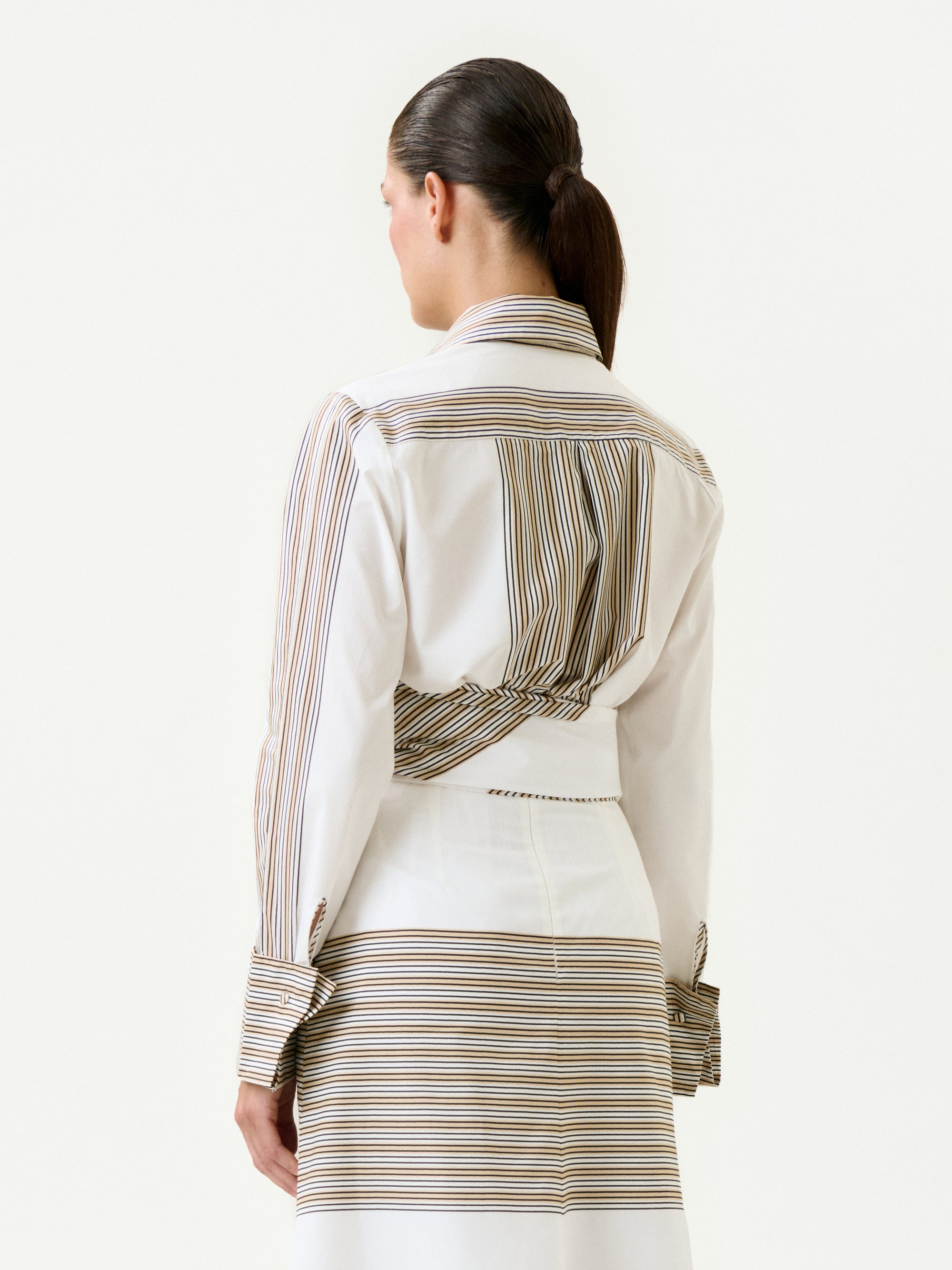 A woman with dark hair in a low ponytail is shown from the back, wearing the 100% cotton Angeles Shirt in soft Italian fabric with stripes and a matching skirt against a plain, light background.