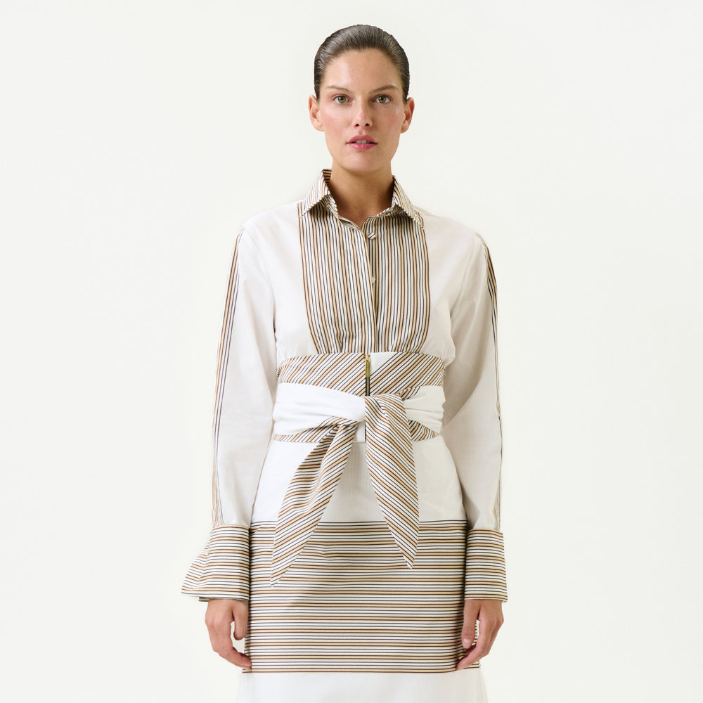 A woman faces forward in the Angeles Shirt, a long-sleeve top made from 100% cotton Italian fabric with beige and brown vertical stripes, matching cuffs, and a wide striped belt at the waist. She wears it over a skirt with horizontal stripes against a plain white background.