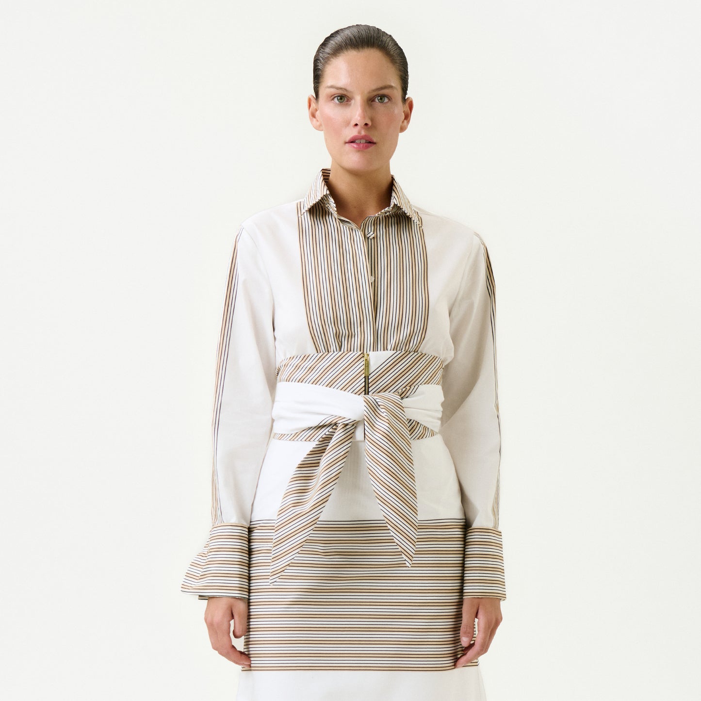 A woman faces forward in the Angeles Shirt, a long-sleeve top made from 100% cotton Italian fabric with beige and brown vertical stripes, matching cuffs, and a wide striped belt at the waist. She wears it over a skirt with horizontal stripes against a plain white background.