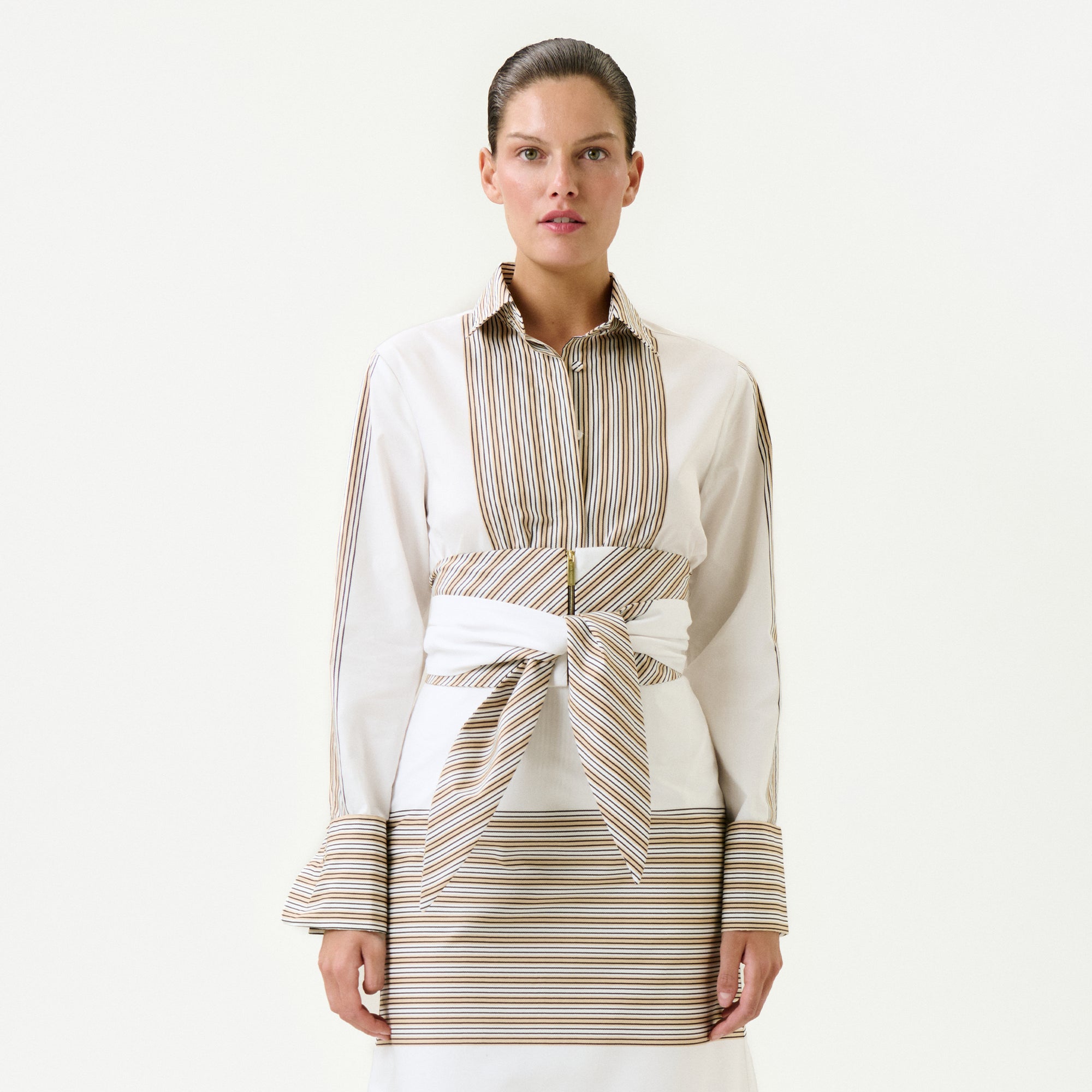 A woman faces forward in the Angeles Shirt, a long-sleeve top made from 100% cotton Italian fabric with beige and brown vertical stripes, matching cuffs, and a wide striped belt at the waist. She wears it over a skirt with horizontal stripes against a plain white background.