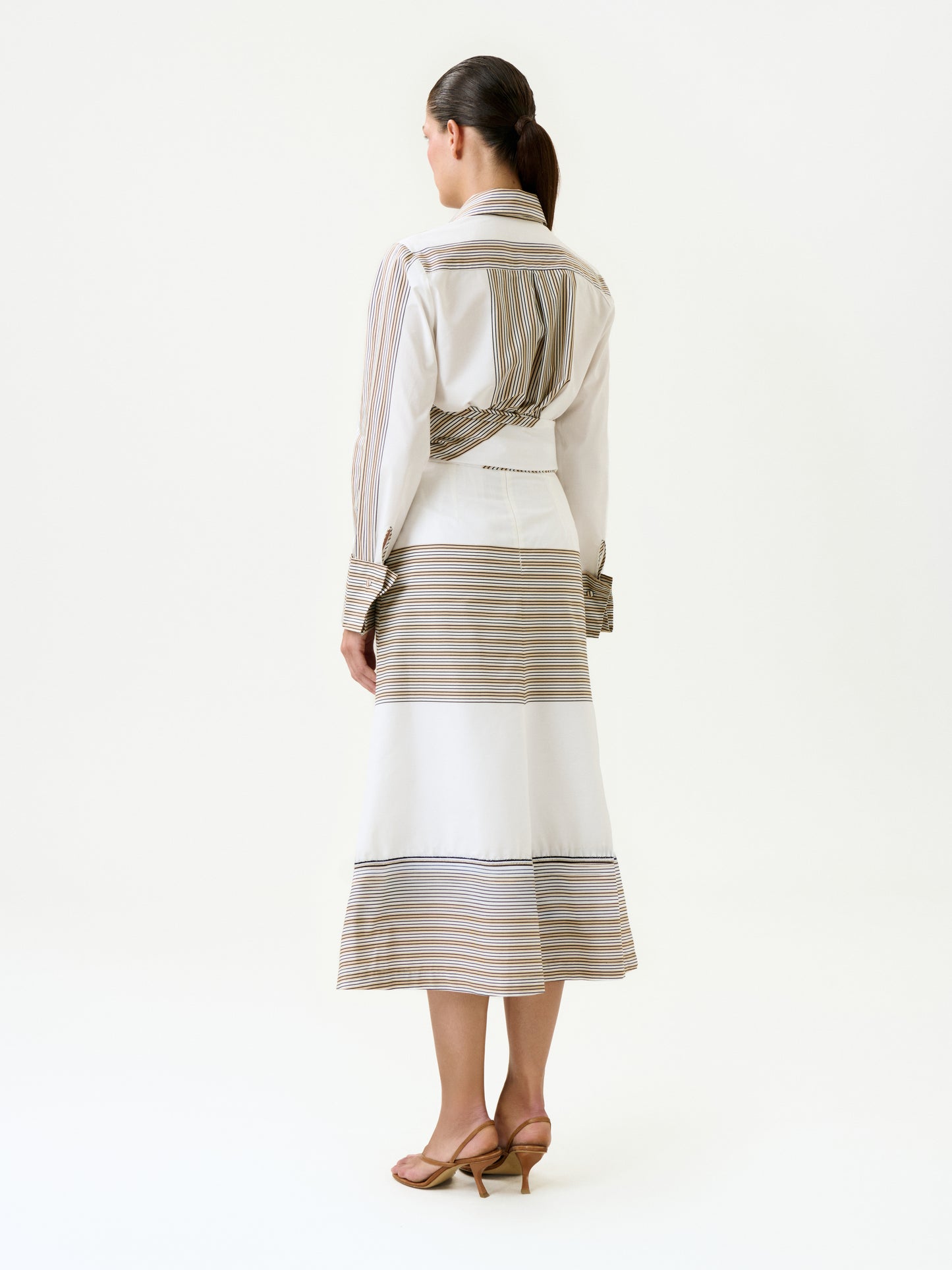 A woman with a ponytail stands facing away, wearing the Lyla Skirt—a long, 100% cotton Italian fabric skirt with horizontal beige and gray stripes—and a matching white long-sleeve shirt, finished with a detachable belt and brown heeled sandals.