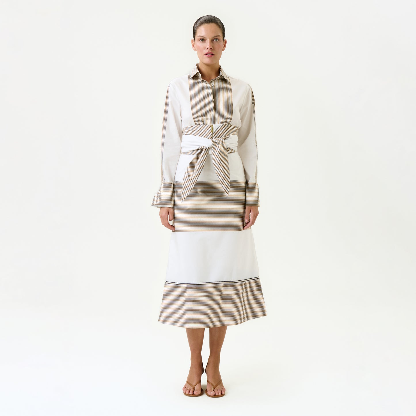 A woman faces forward against a plain white background, wearing the Lyla Skirt—a long, Italian-fabric skirt with beige and white horizontal stripes—paired with a matching collared top and a wide detachable belt at her waist. She completes the look with sandals.