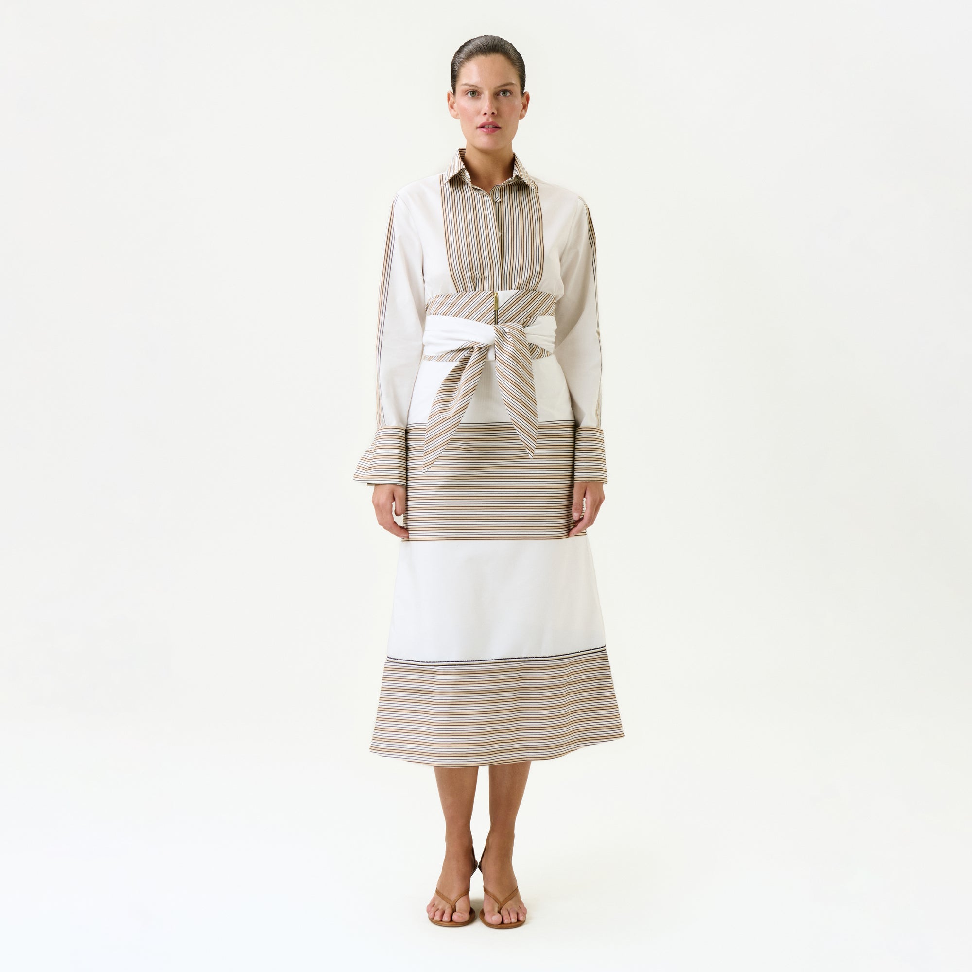 A woman faces forward against a plain white background, wearing the Lyla Skirt—a long, Italian-fabric skirt with beige and white horizontal stripes—paired with a matching collared top and a wide detachable belt at her waist. She completes the look with sandals.