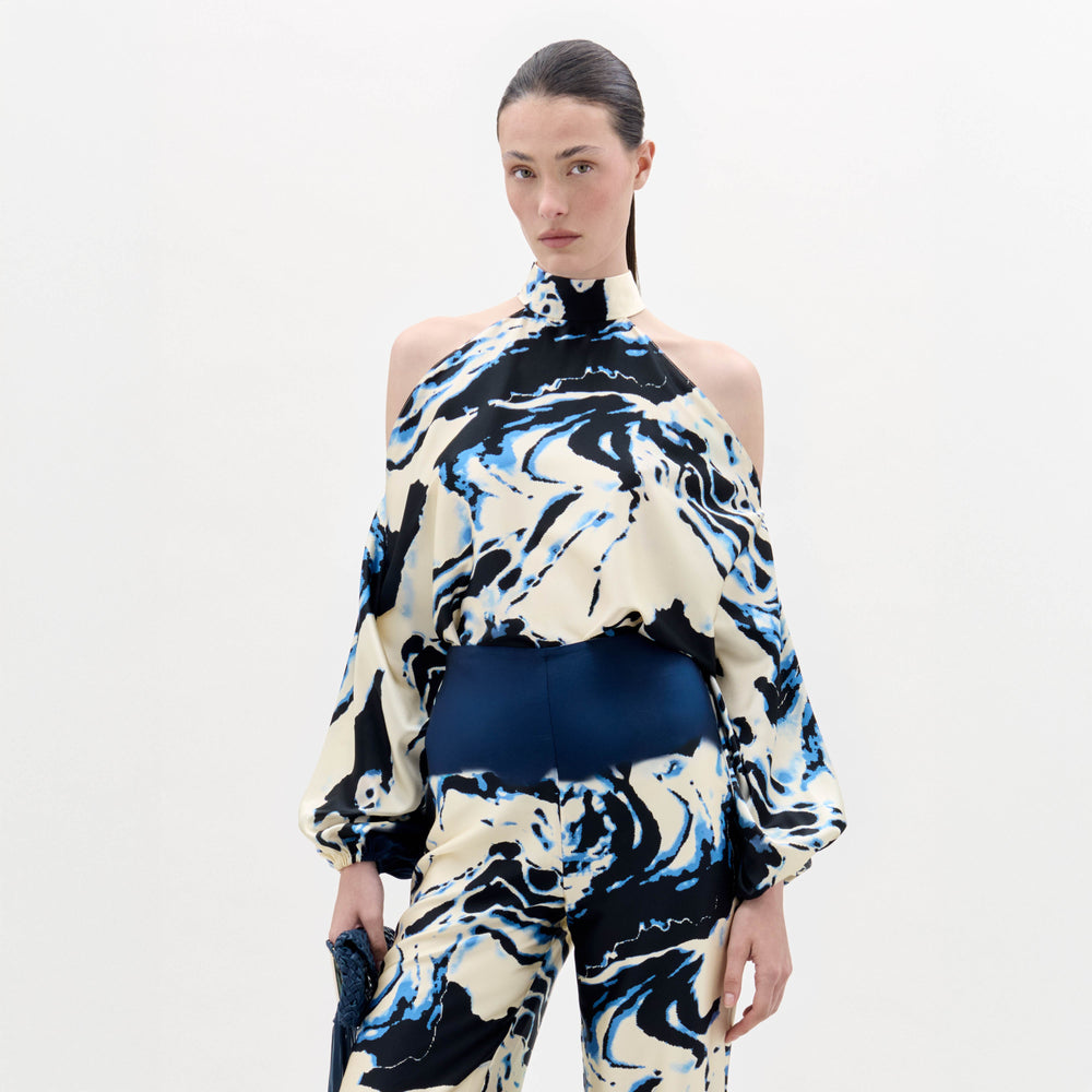 A woman stands against a plain background wearing the Safira Blouse, a high-neck, cold-shoulder top with matching wide-leg pants in a blue, black, and white marble pattern.