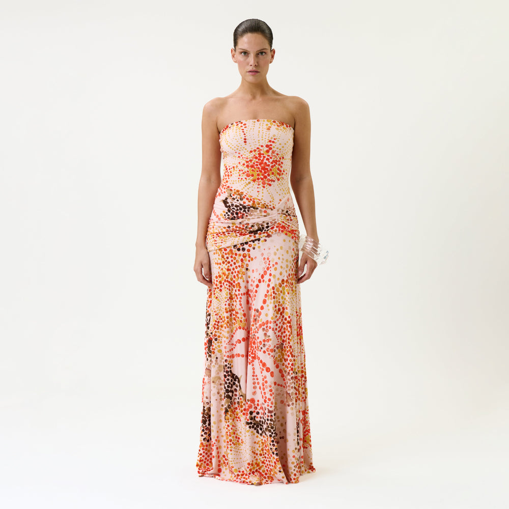 A woman stands against a plain white backdrop wearing the Shani Dress, a strapless, floor-length gown featuring a colorful abstract dot pattern in shades of orange, pink, and brown on a white background.