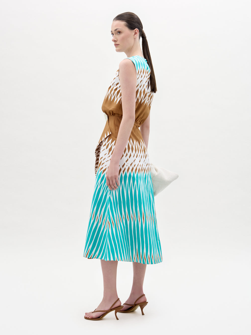 A woman stands sideways against a white background wearing the Kendall Dress—a sleeveless, fit-and-flare silhouette in brown, white, and turquoise. She holds a white clutch, has straight dark hair tied back, and wears brown heeled sandals.