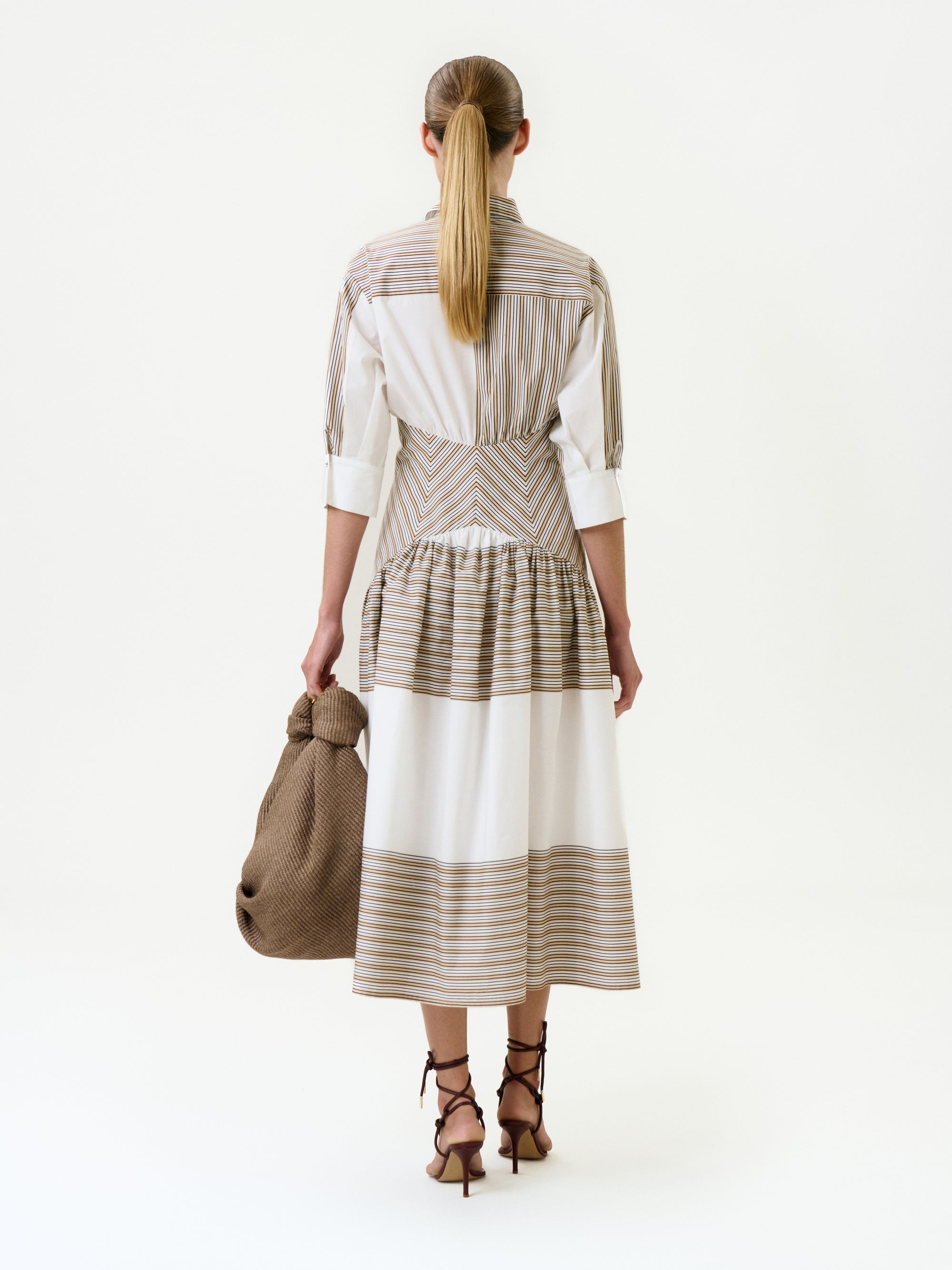A woman faces away, wearing the Indy Dress—a 100% cotton midi with beige stripes, three-quarter sleeves, and a gathered skirt. Her hair is in a low ponytail, and she accessorizes with a textured brown bag and brown lace-up heels.