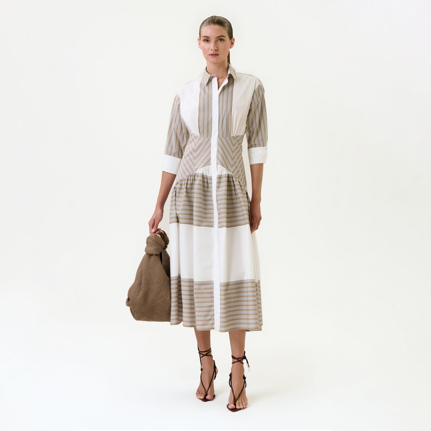 A woman stands against a white background wearing the Indy Dress—a long, collared midi dress with beige and white stripes in 100% Italian cotton. She carries a large brown textured bag and pairs the look with black strappy sandals.