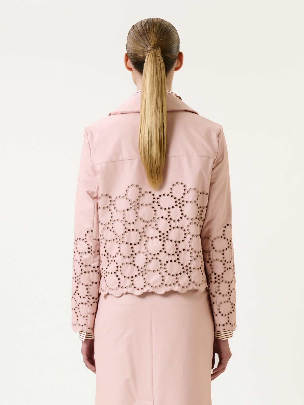 A woman with long, straight blonde hair in a ponytail is seen from the back against a plain white background, wearing the Belen Jacket—a light pink piece with eyelet detailing—paired with a matching pink skirt.