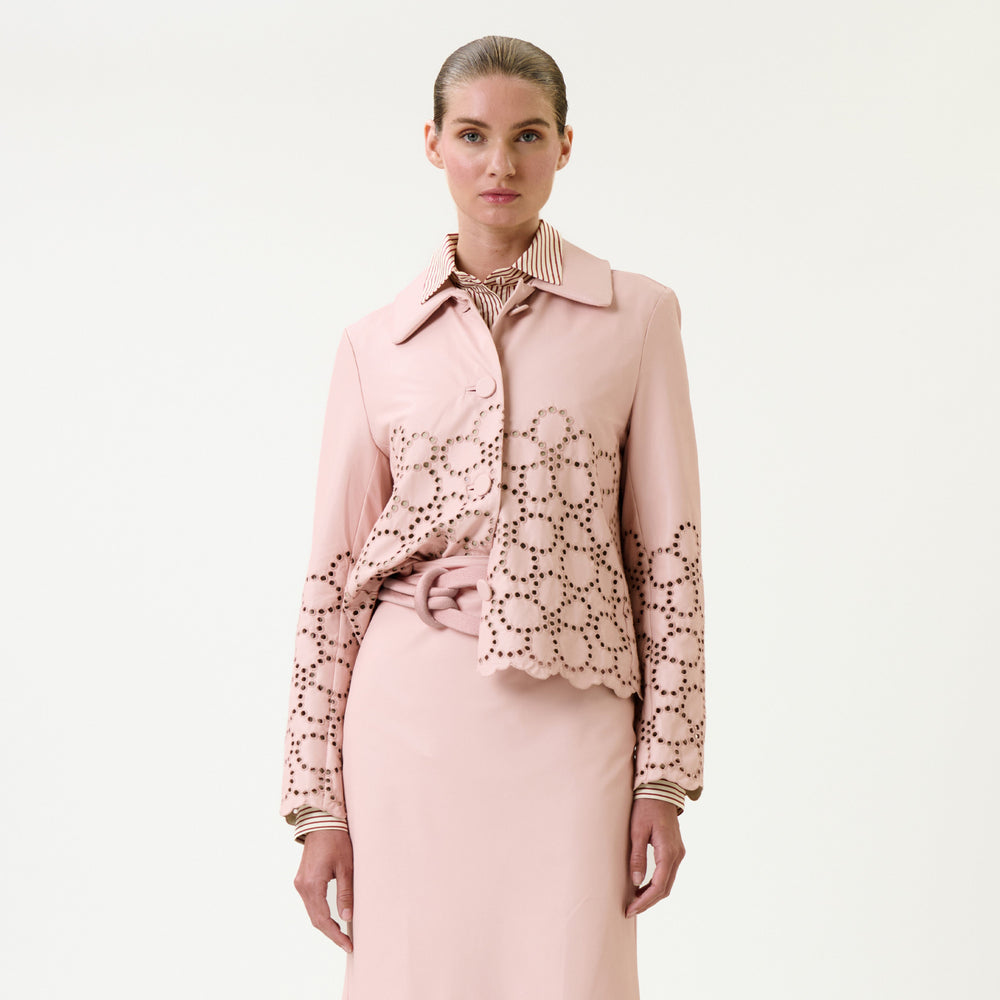 A woman stands against a plain background, wearing the Belen Jacket—a light pink piece featuring circular cut-out patterns on the sleeves and lower half—paired with a matching skirt over a striped collared shirt. Her hair is slicked back.