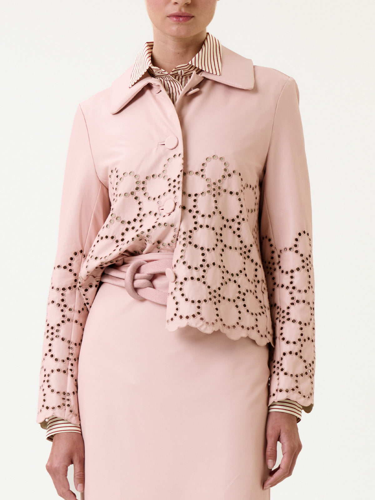 A woman wearing the Belen Jacket in light pink, featuring laser-cut floral patterns, scalloped edges, and a belted waist, layered over a matching skirt and striped collared shirt. Only her lower face is visible.