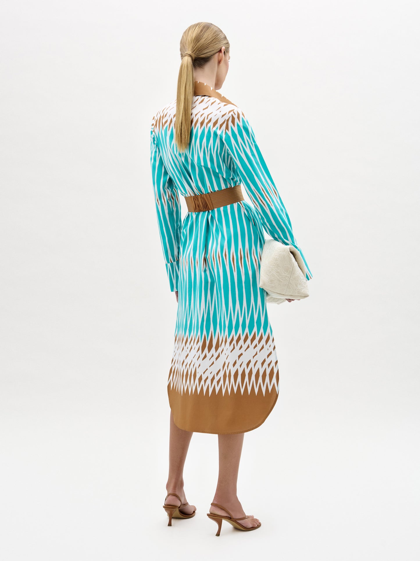 A woman with blonde hair in a low ponytail stands facing away, wearing the Ronzone Dress—a teal, white, and brown patterned Italian-fabric midi with a detachable brown belt—tan heeled sandals, and holding a white clutch.