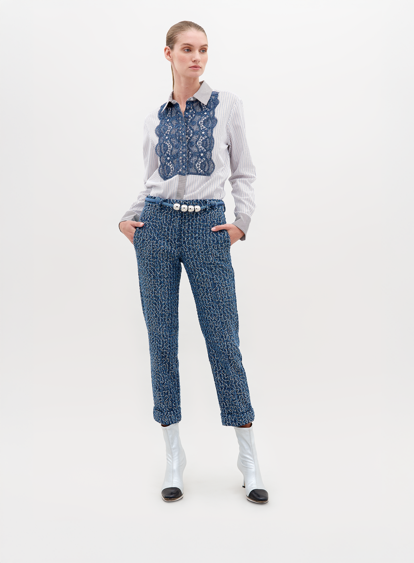 A woman stands against a plain background wearing a white blouse with blue lace detail, Garmet Pant Navy Sequins, a pearl belt, and white ankle boots with black tips. This stylish ensemble is available for pre-order now and will ship by November 15th, 2024.