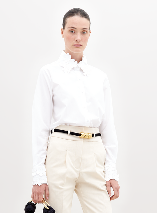 A person with dark hair tied back, wearing a Flower Blouse White with flower details on the collar and sleeves, beige pants, and a black belt with gold accents, holding a small black handbag. Pre-order now to ensure your outfit ships by November 15th, 2024.