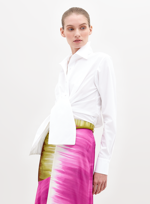 A person wearing a Mercury Blouse White and a vibrant pink skirt with green accents, standing against a plain white background, available for pre-order now. Ships by November 15th, 2024.