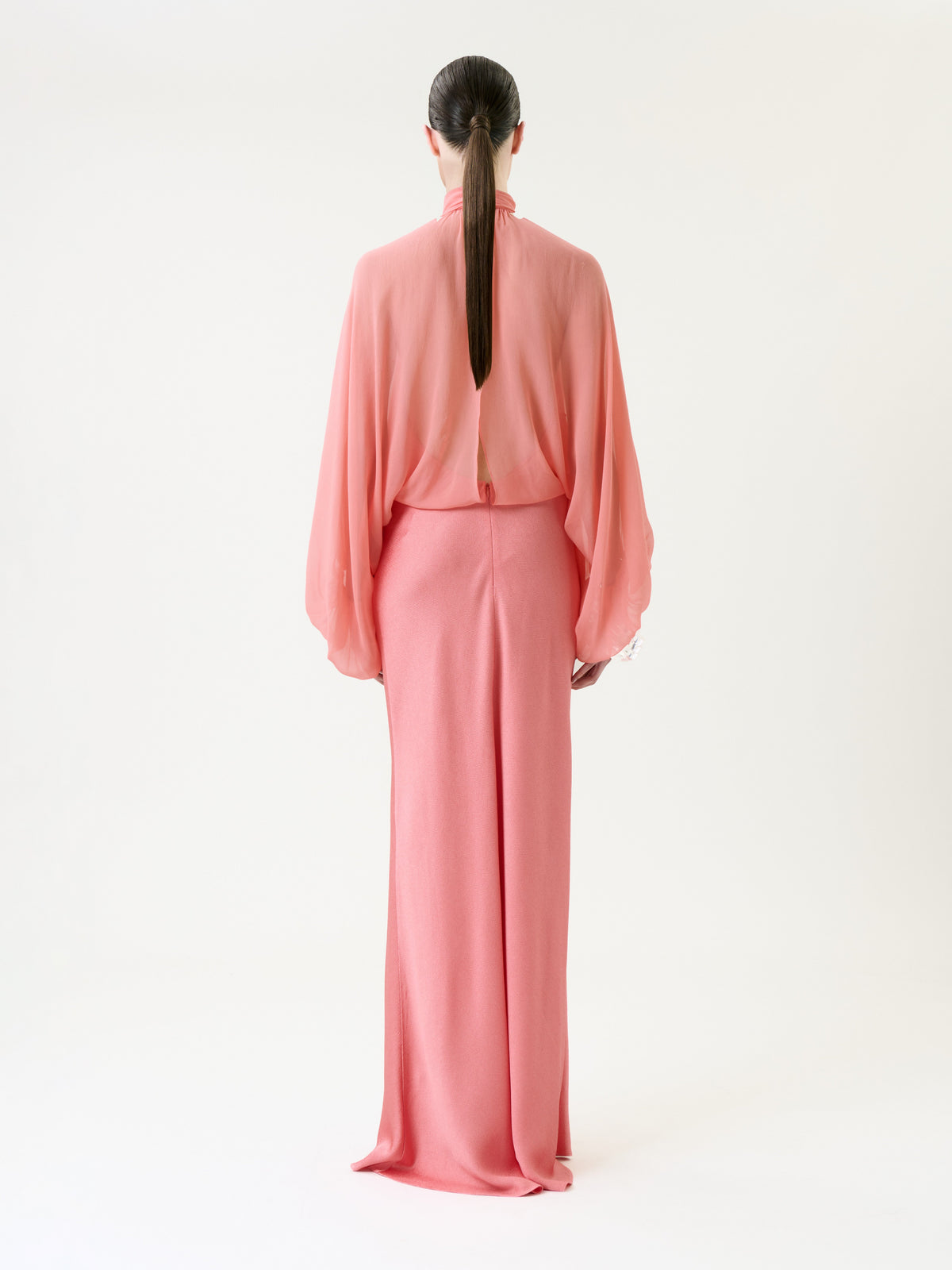 A person with long, straight brown hair stands facing away, wearing the Martine Dress—a sheer pink blouse paired with a long pink skirt—against a plain white background.