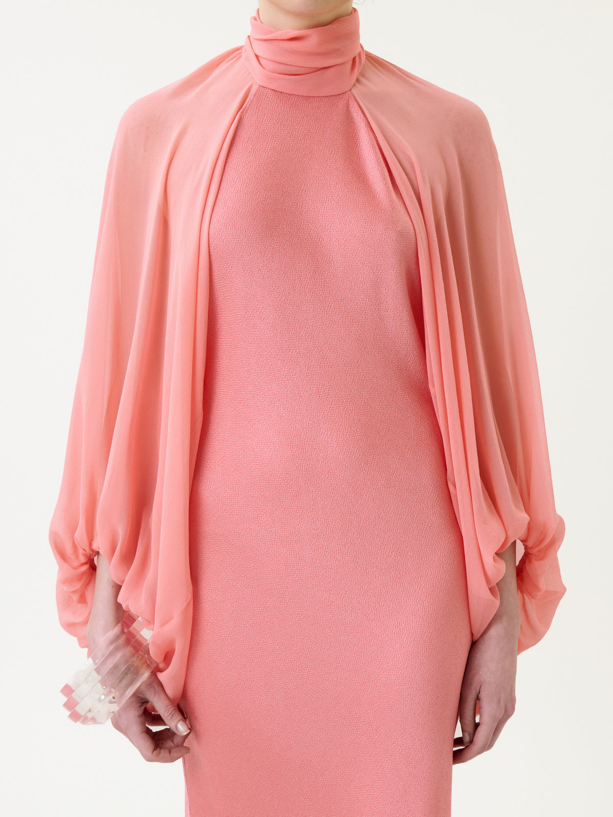 A person wearing the Martine Dress—a high-neck, sleeveless, textured pink dress—styled with a sheer, flowing pink cape and holding a translucent clutch.