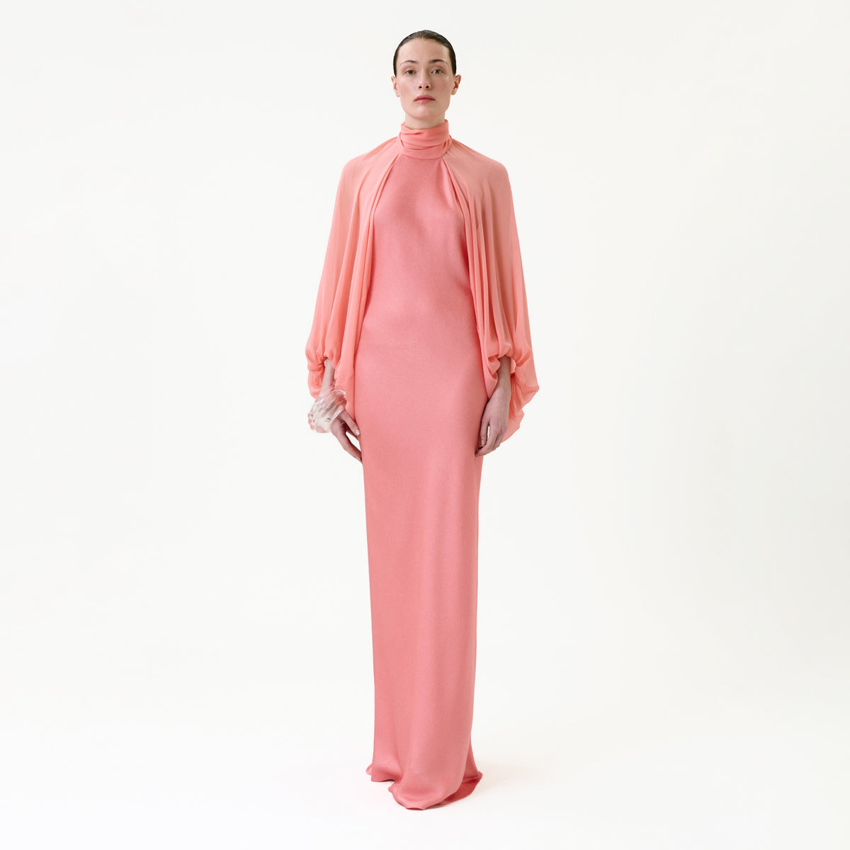 A person stands against a white background wearing the Martine Dress, a long pink high-neck style with flowing sleeves, holding a small pink clutch.
