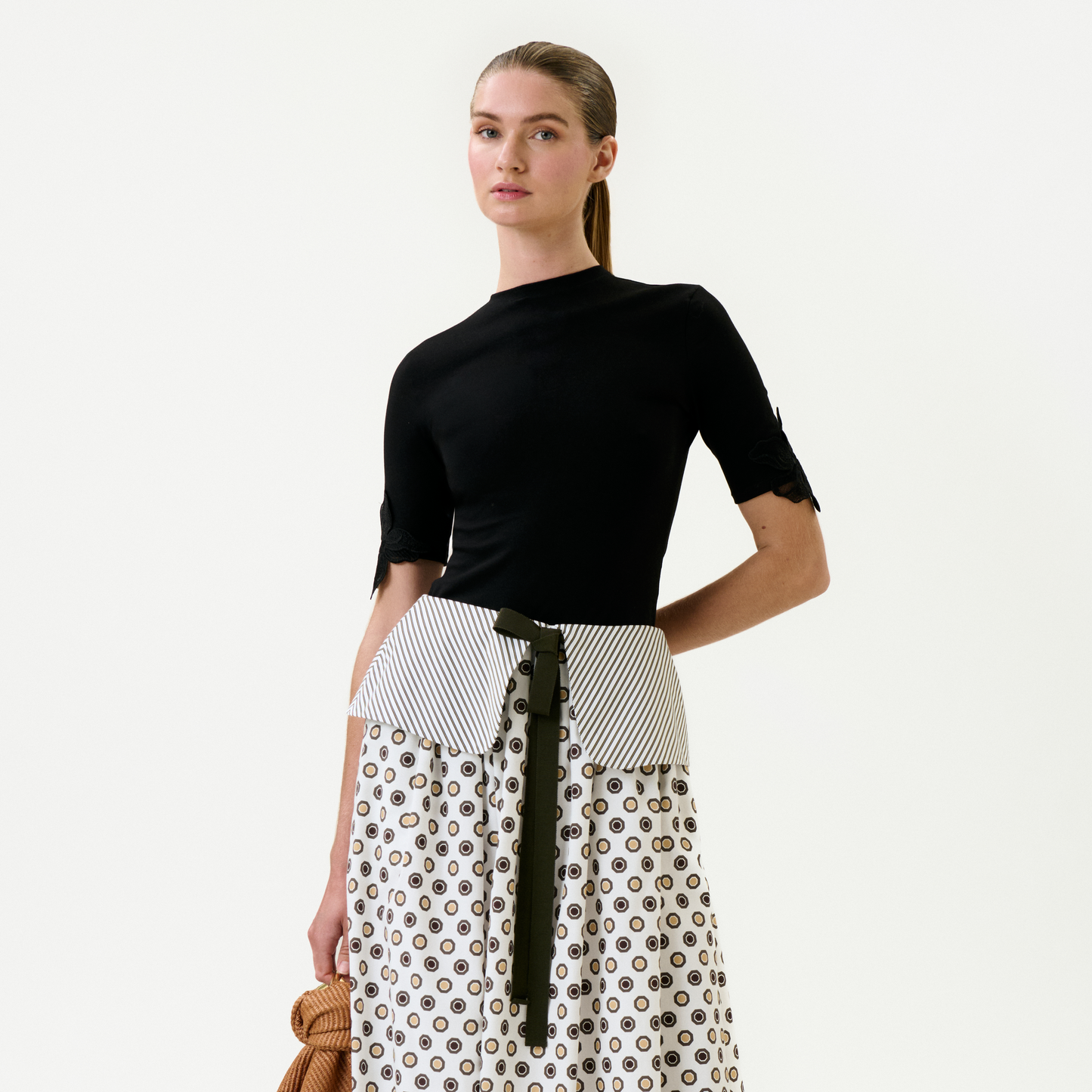 A woman poses against a plain background, wearing the Walda T-Shirt—a fitted black top with elbow-length sleeves—paired with a white patterned Italian fabric skirt, striped waistband, black belt, and holding a brown textured item in her left hand.
