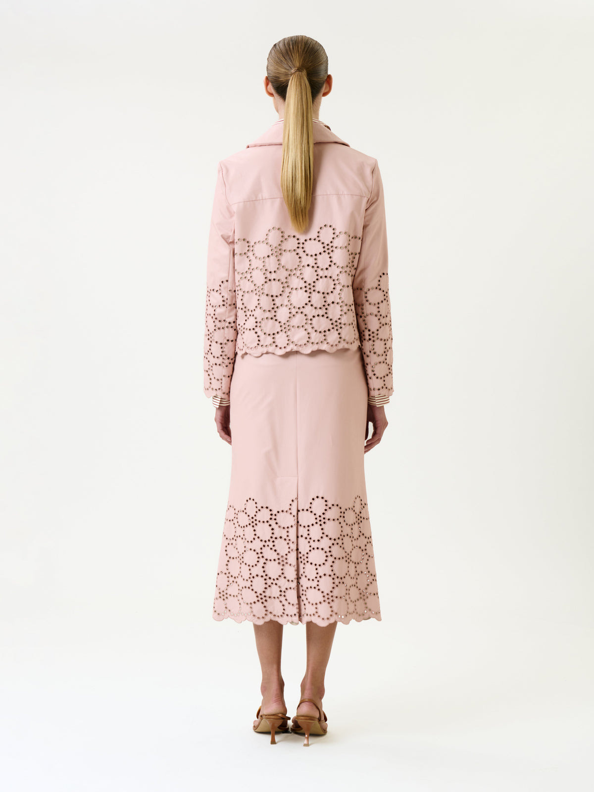 A woman stands facing away, wearing a light pink jacket and the Atira Skirt, both featuring decorative cut-out flower patterns along the hem and sleeves. She has long straight hair tied back and is wearing heeled sandals.