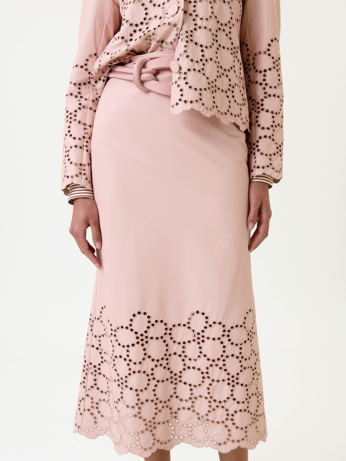A person wearing the Atira Skirt in light pink, featuring circular cutout patterns near the hem, paired with a matching jacket. Their hands rest by their sides against a plain white background.