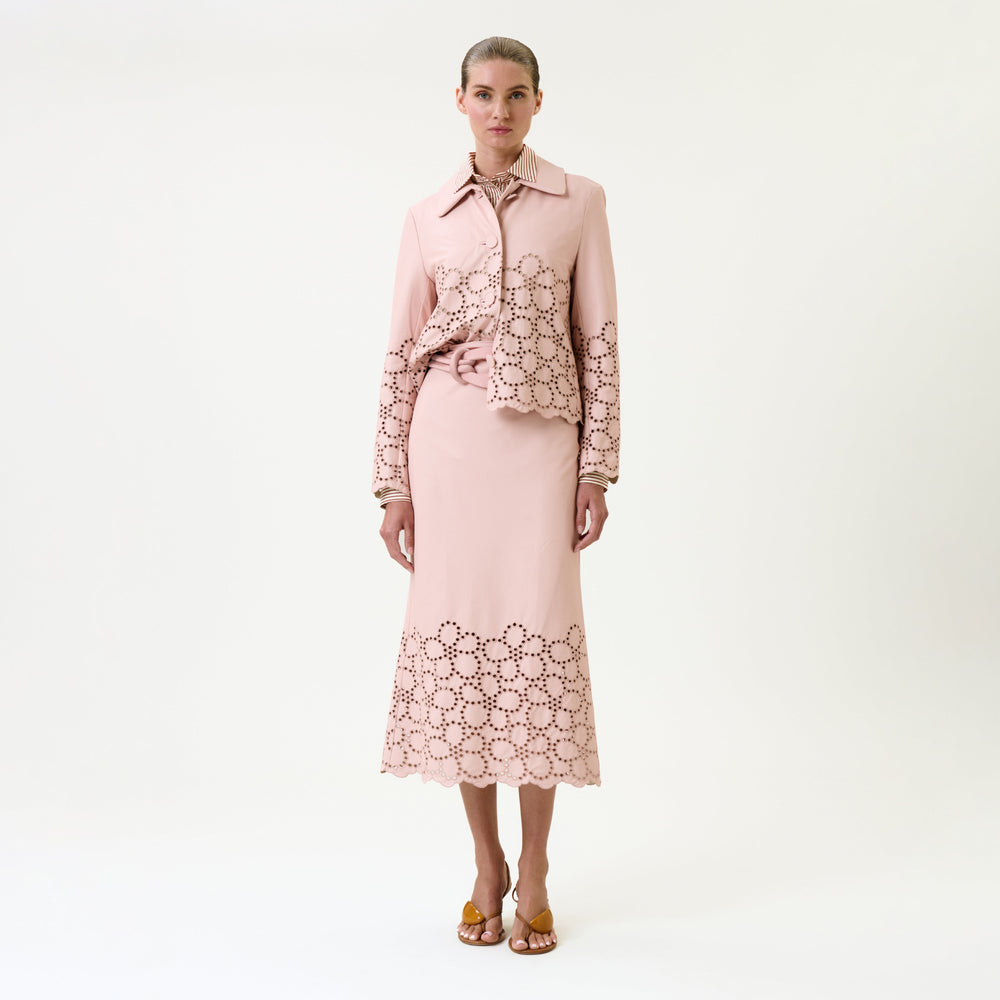 A woman stands against a white background wearing the Atira Skirt and a matching light pink shirt with laser-cut floral patterns, paired with yellow heeled sandals.