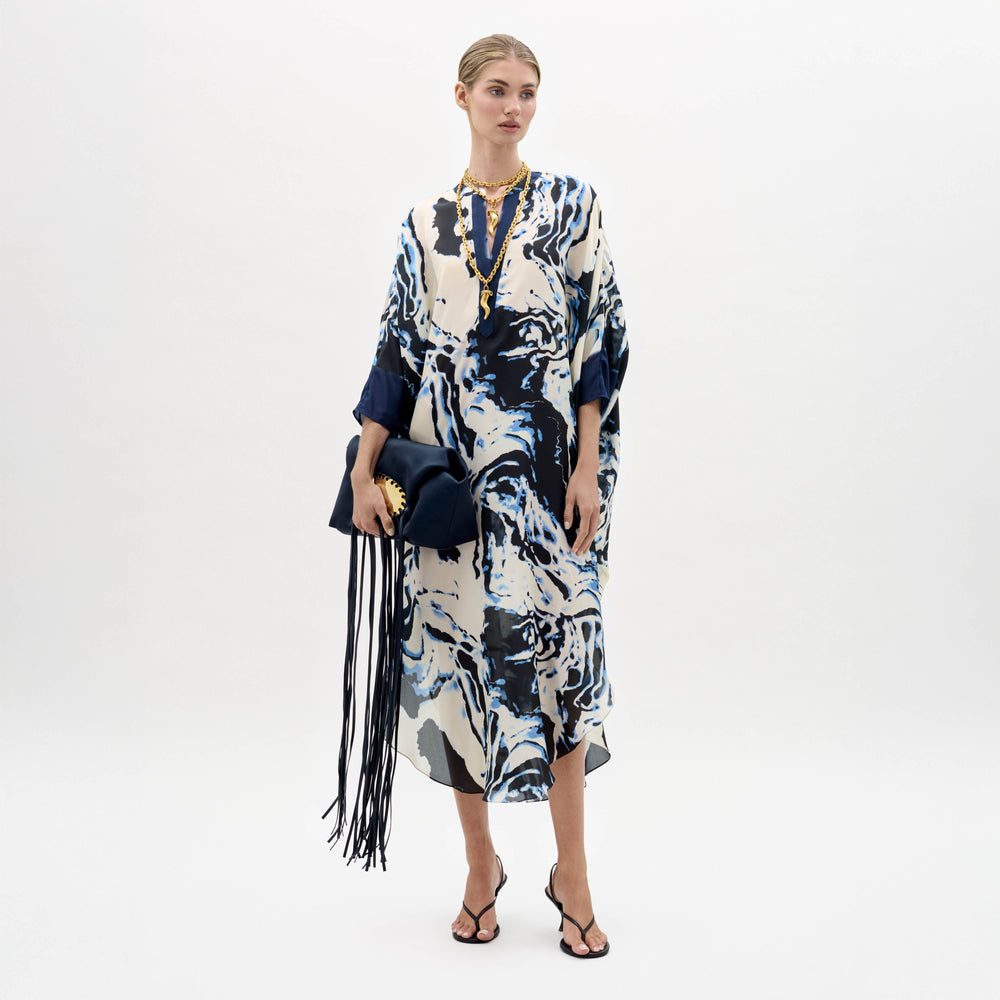 A woman stands against a plain background wearing the Elea Tunic with an abstract pattern, black sandals, gold jewelry, and holding a navy fringe bag.