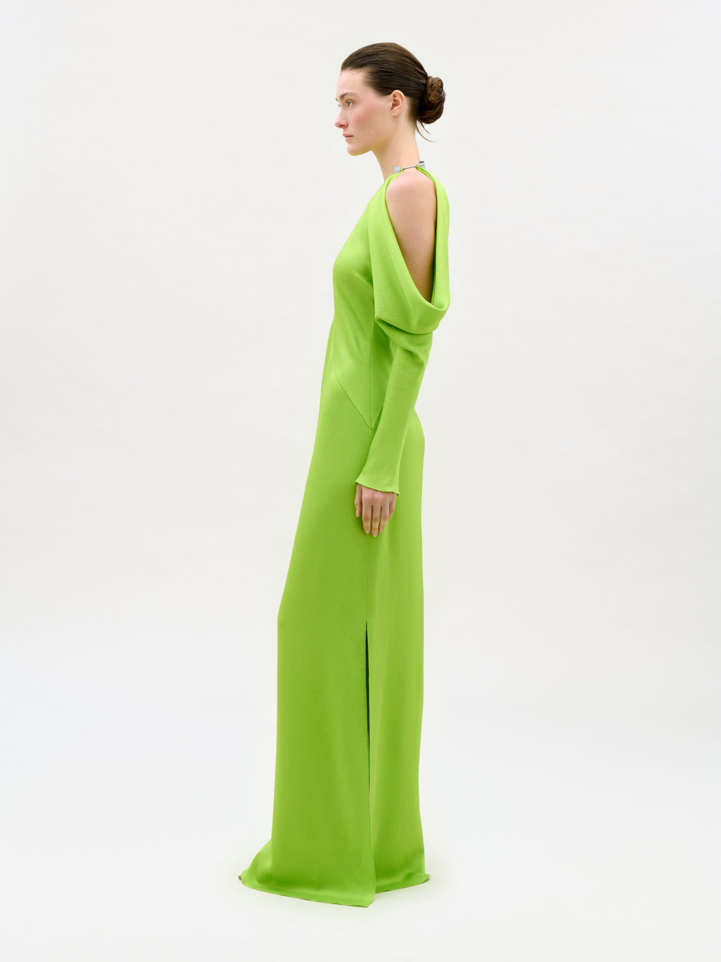 A woman with brown hair in a low bun stands in profile against a plain white background, wearing the bright green Antonia Dress—a column silhouette maxi with open shoulders and long sleeves.