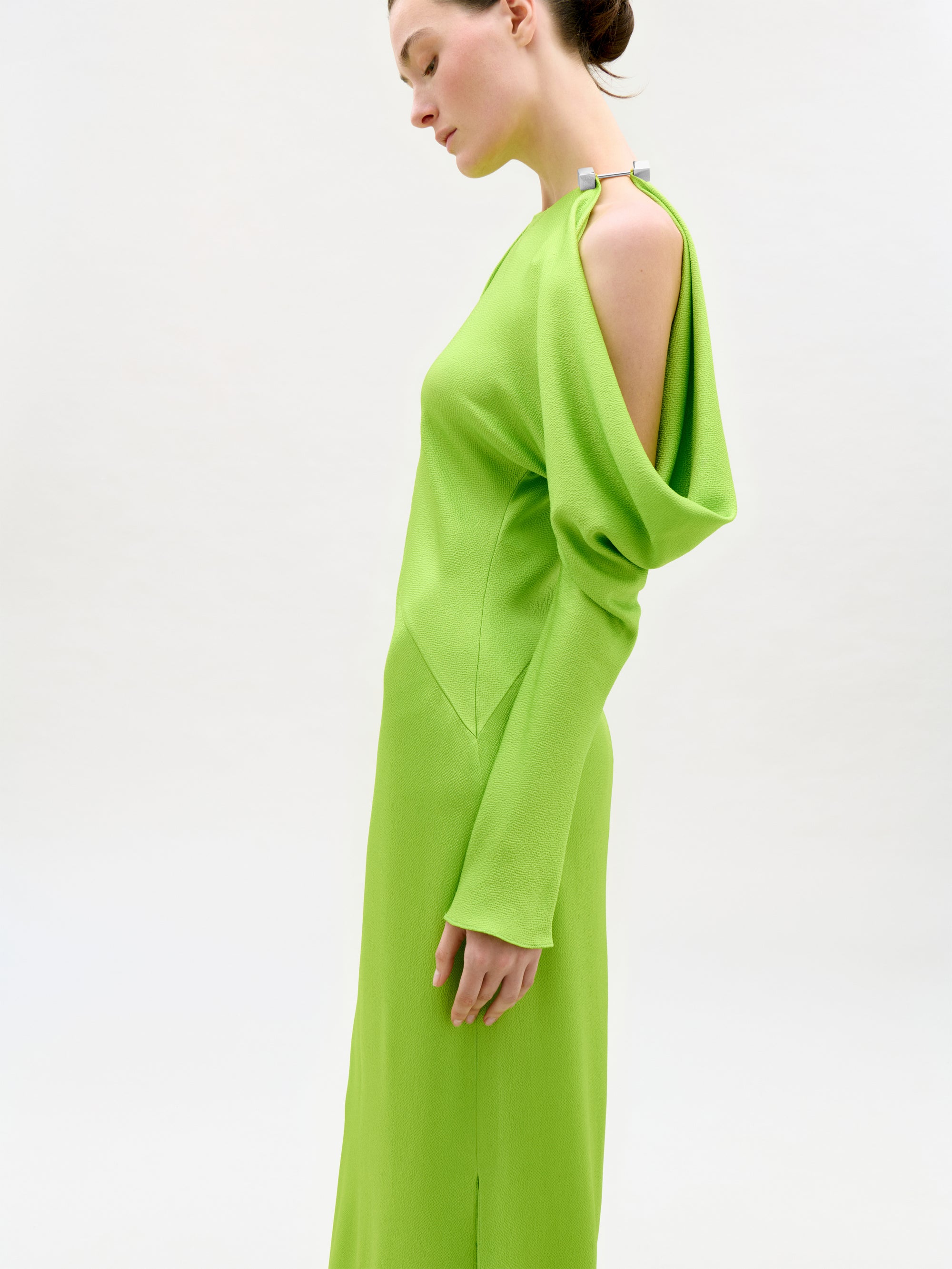 A woman stands in profile against a plain light background, wearing the Antonia Dress—a bright green Italian fabric maxi with open shoulder, draped sleeve detail, and an elegant column silhouette.