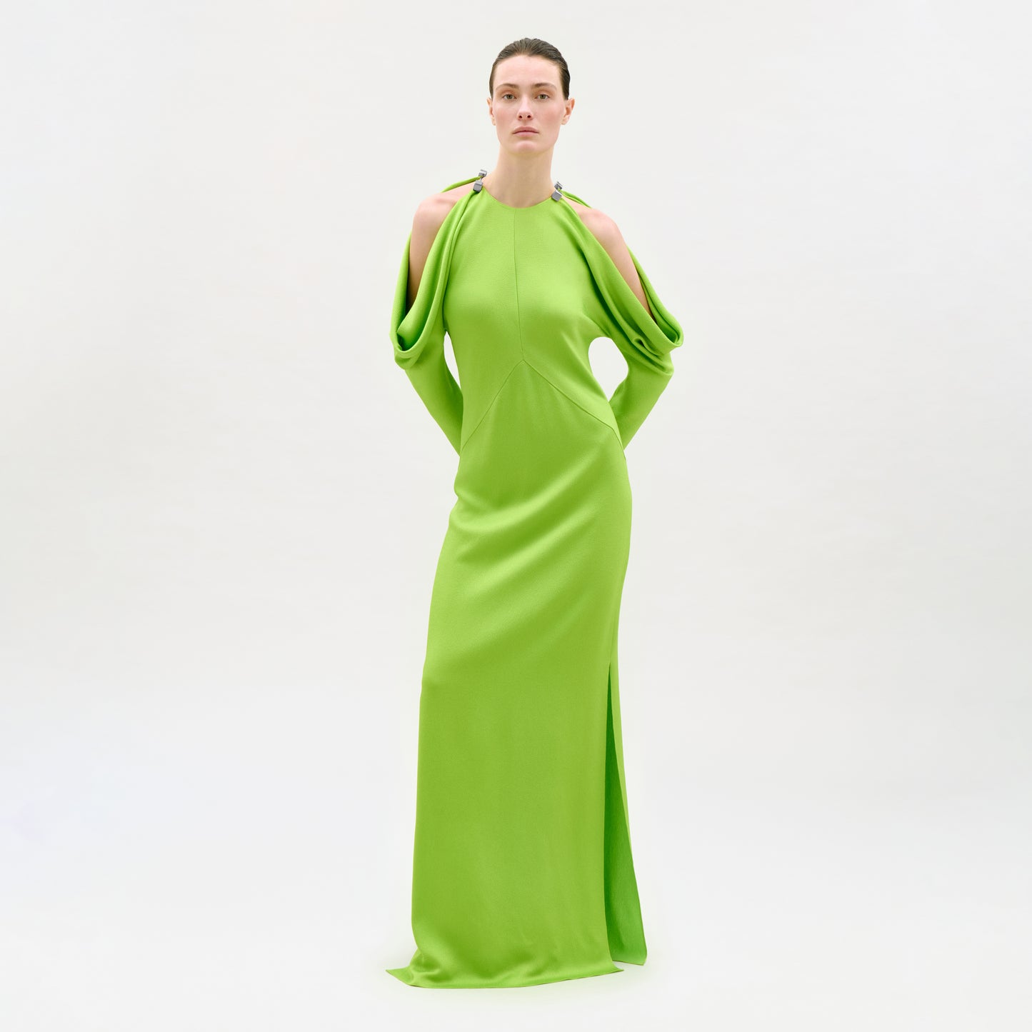A person poses before a white backdrop in the Antonia Dress, a bright green Italian maxi with dramatic draped sleeves, shoulder cutouts, high neckline, and a straight column silhouette.