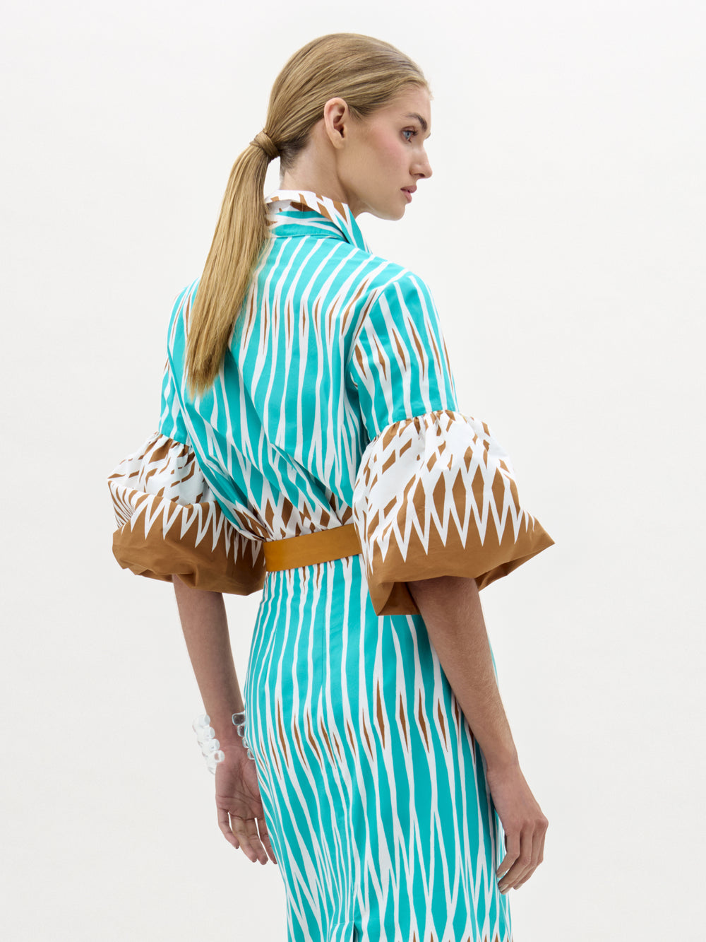 A woman with blonde hair in a ponytail models the Helina Blouse—a turquoise and brown patterned top with wide bell sleeves—standing against a plain light background, facing away with her head turned to the side.