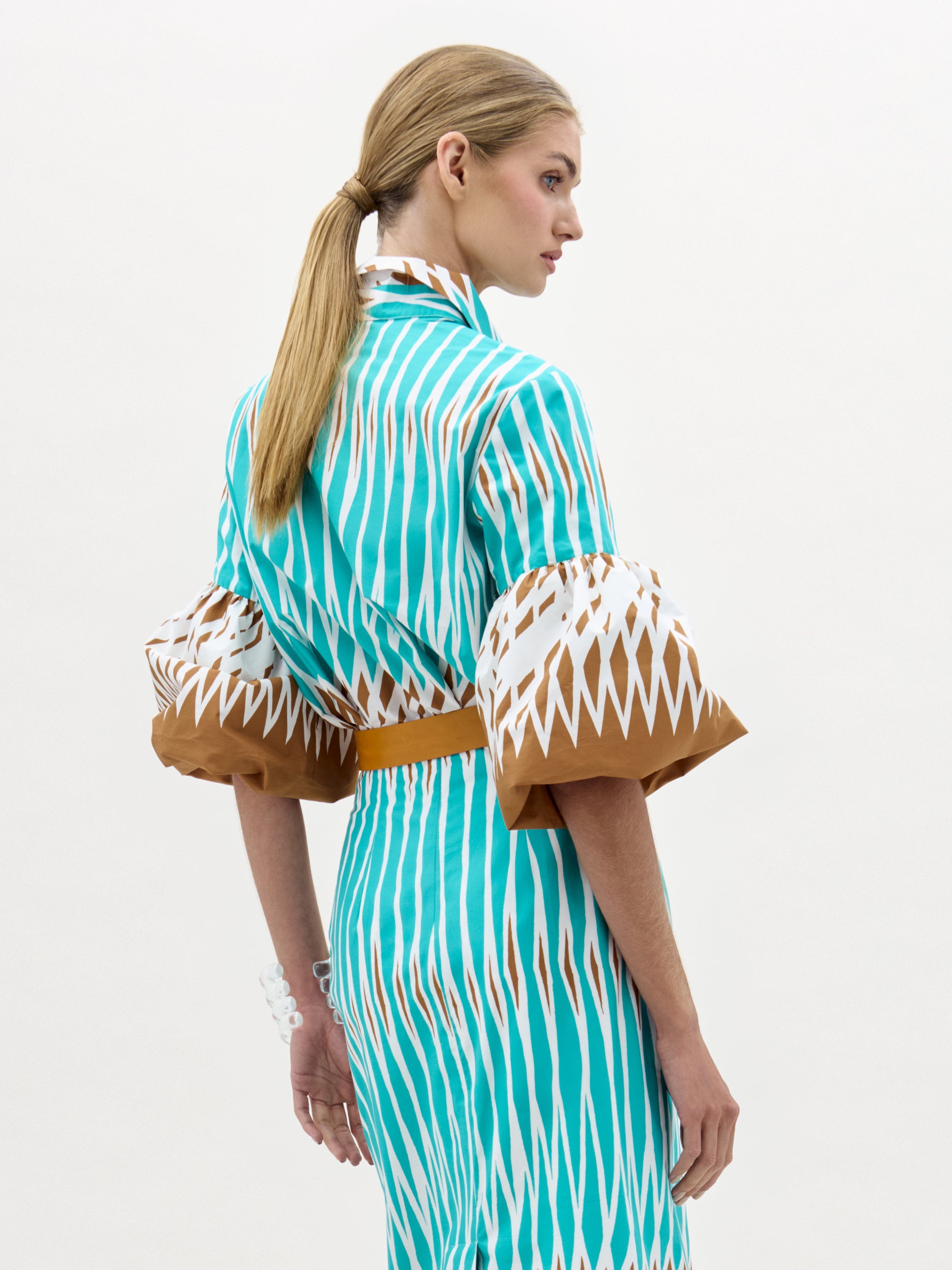 A woman with blonde hair in a ponytail models the Helina Blouse—a turquoise and brown patterned top with wide bell sleeves—standing against a plain light background, facing away with her head turned to the side.