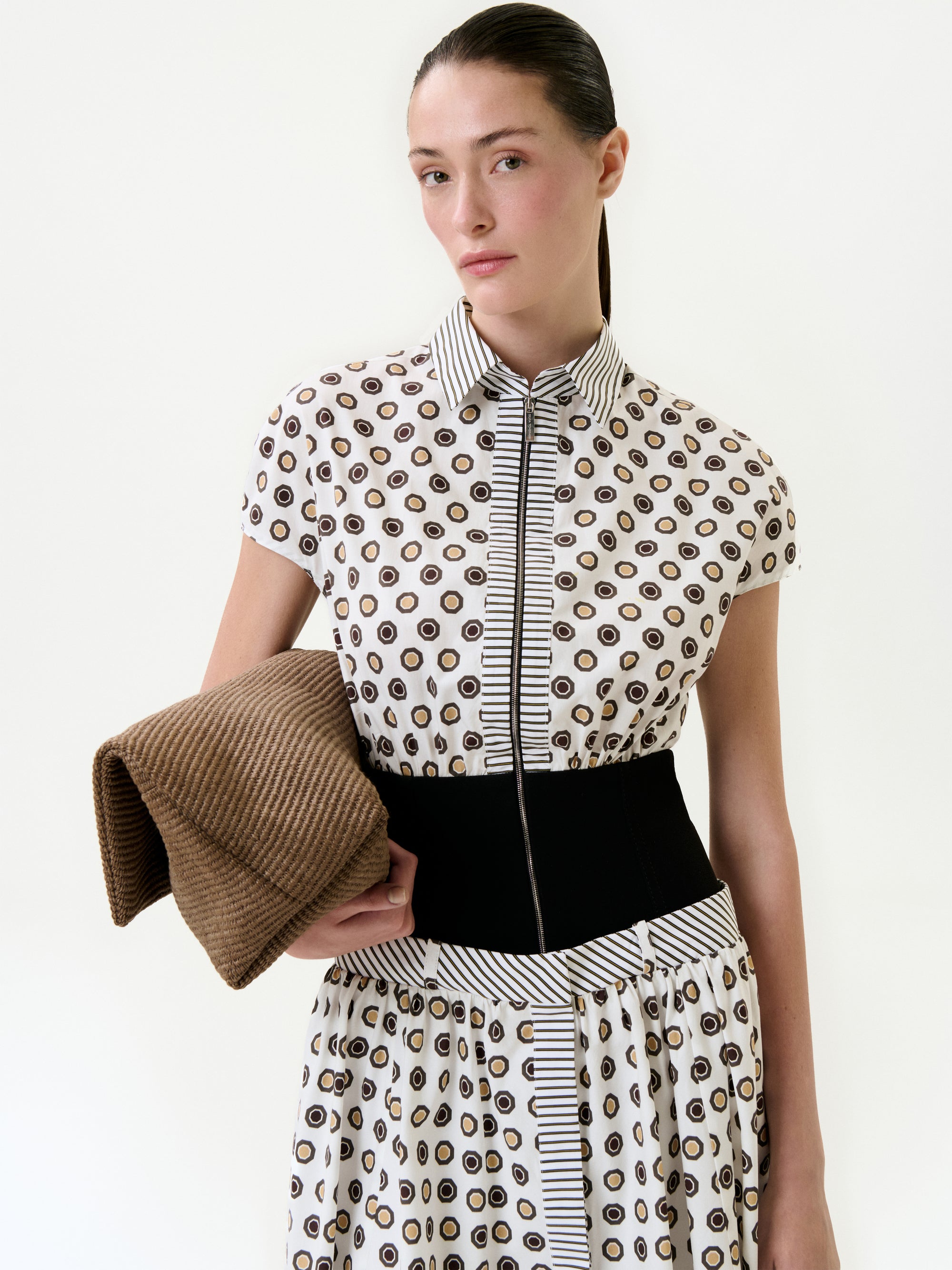 A woman with straight brown hair pulled back wears the Odalis Dress—a patterned 100% cotton short-sleeve midi—with a wide black belt. She holds folded brown textured fabric, standing against a plain white background.