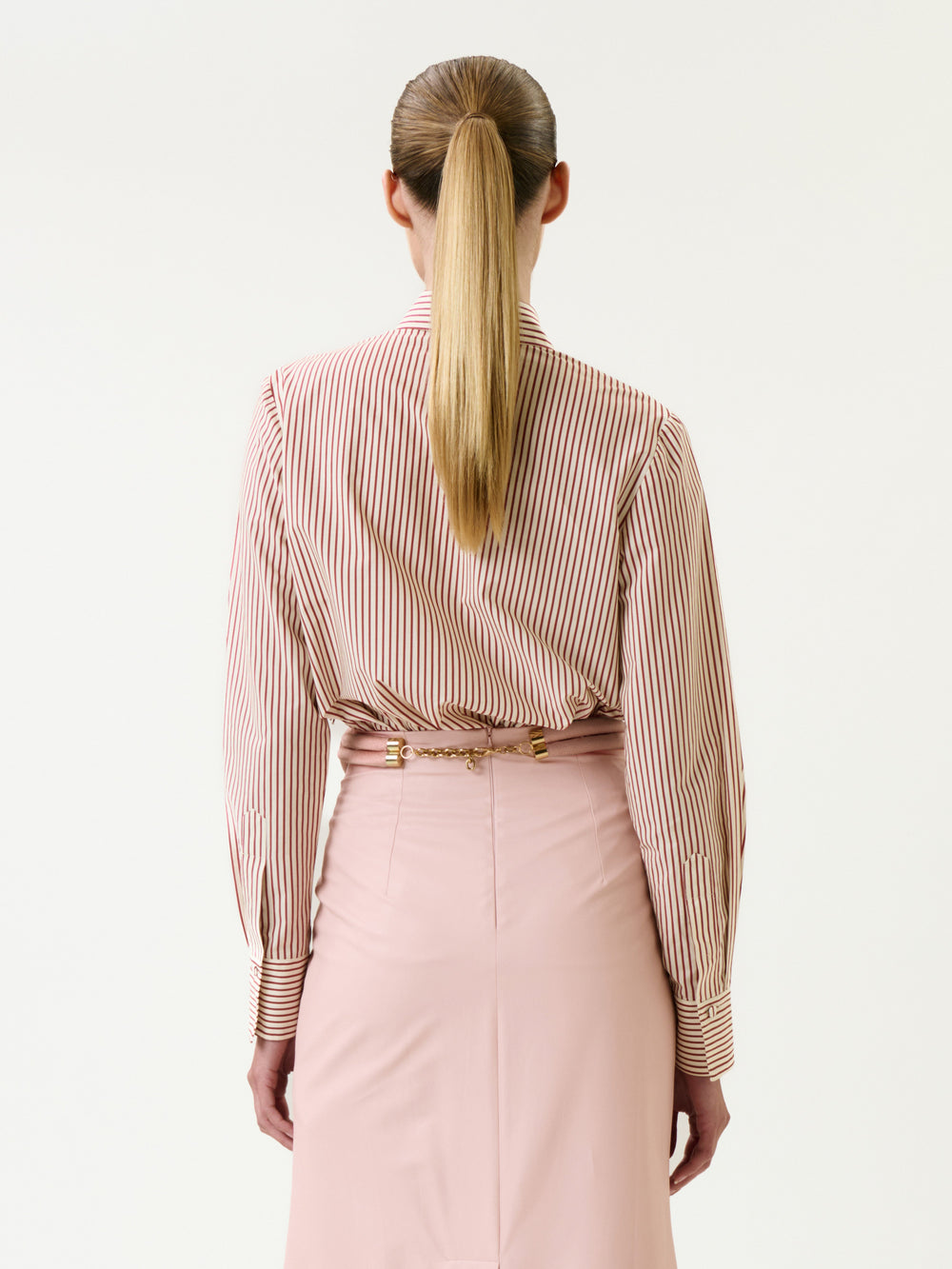 A woman with long blonde hair in a ponytail is seen from behind against a plain white background, wearing the Naroa Shirt—red and white striped—tucked into a high-waisted light pink skirt.