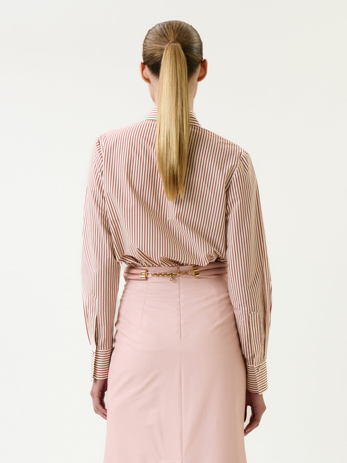 A woman with long blonde hair in a ponytail is seen from behind against a plain white background, wearing the Naroa Shirt—red and white striped—tucked into a high-waisted light pink skirt.