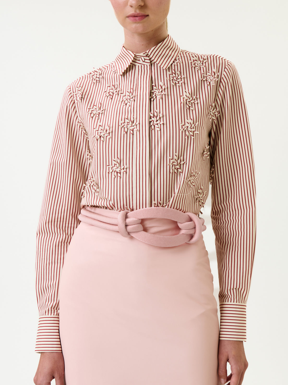A woman wears the Naroa Shirt, a pink and red vertically striped blouse with embroidered details, tucked into a light pink skirt with a large, textured pink belt. Her head is partially cropped out of the image.