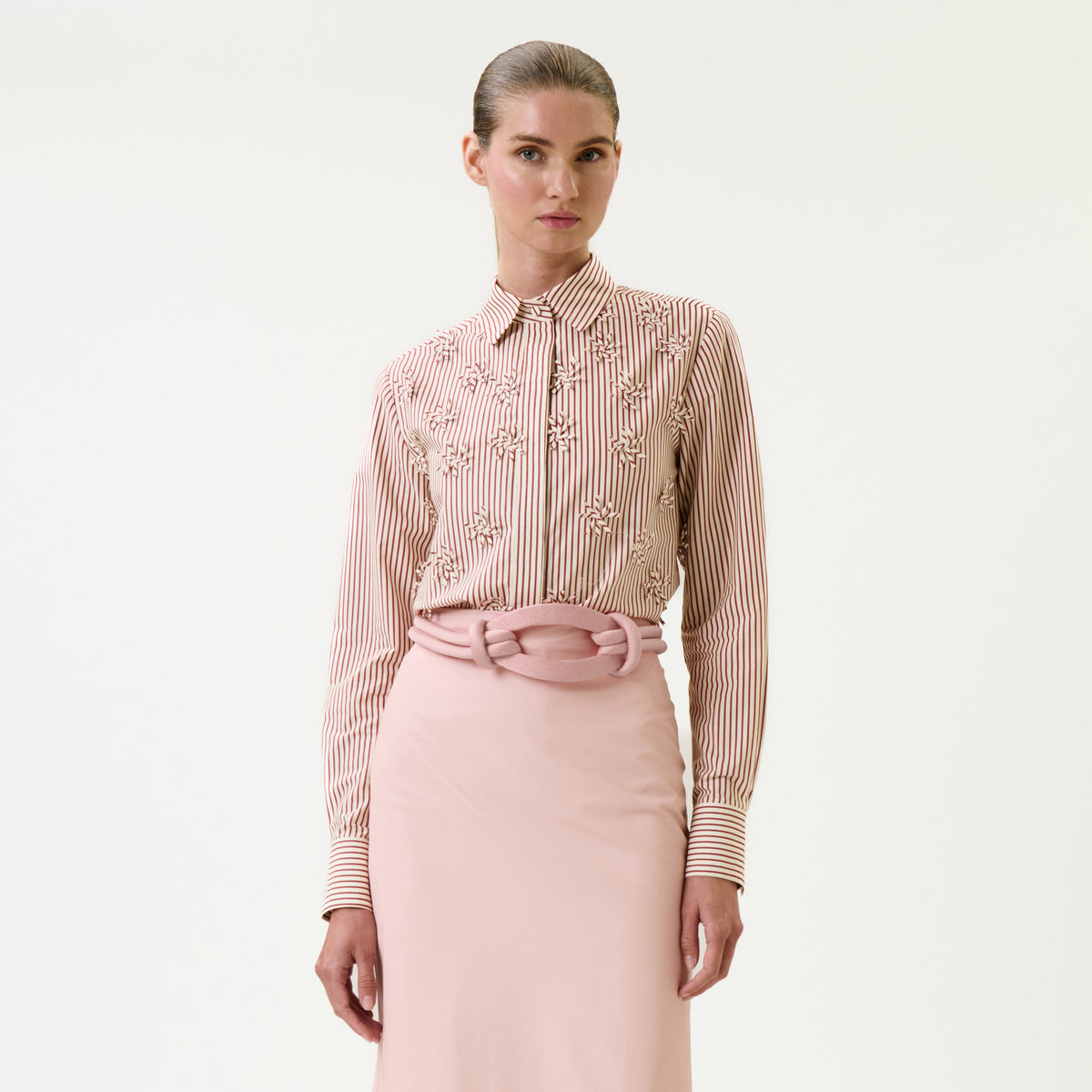 A woman wearing the Naroa Shirt, a pink and white striped blouse with embroidered details, paired with a pale pink skirt and standing against a plain white background.