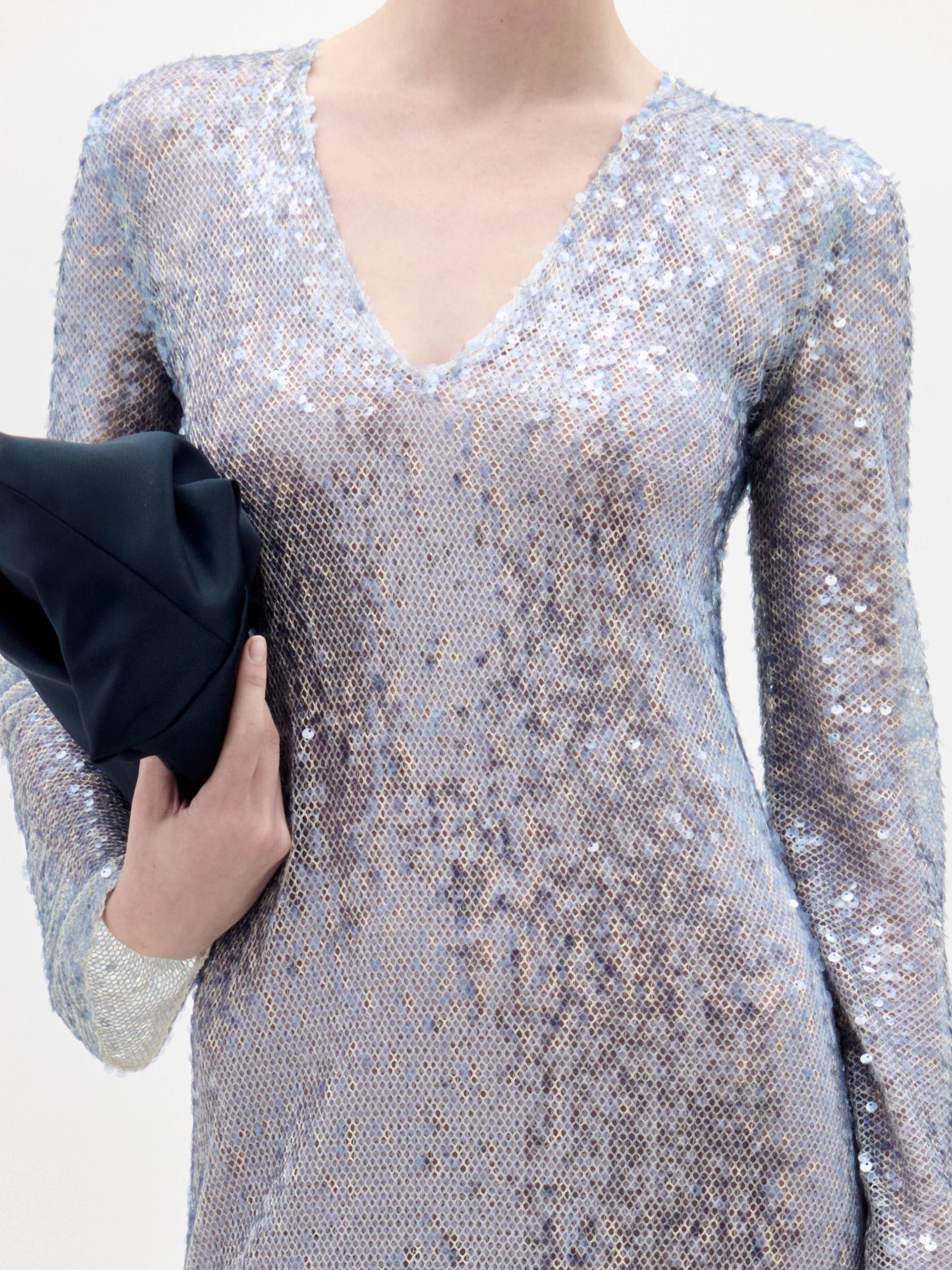 A woman stands against a plain background wearing the Nico Dress, a long-sleeved, floor-length silver sequin gown, and holding a black clutch.