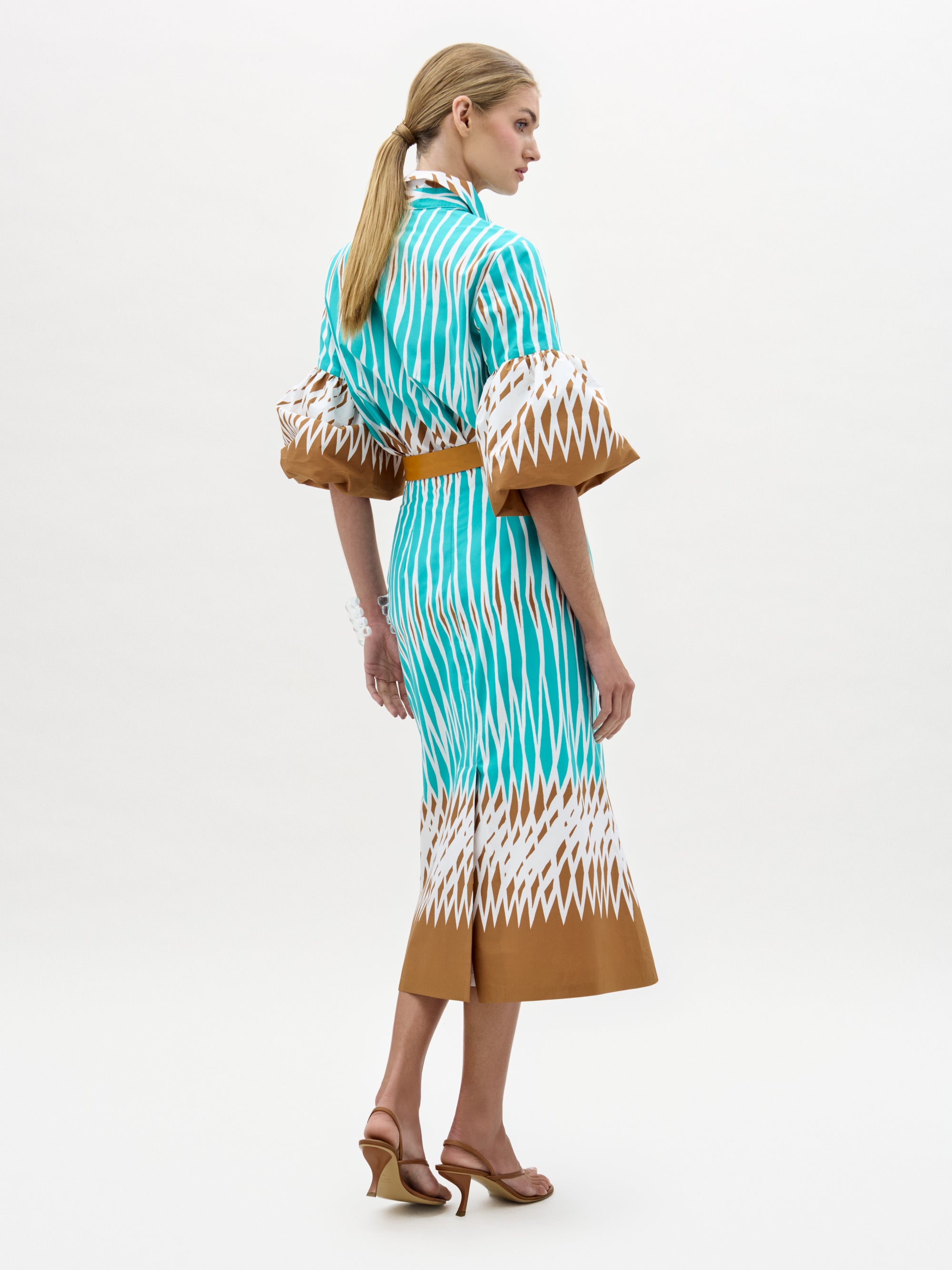 A woman faces away, wearing the Atira Skirt—a high-waisted, teal, white, and brown patterned midi skirt with a brown belt. She pairs it with heels and has long blonde hair tied back against a plain white background.