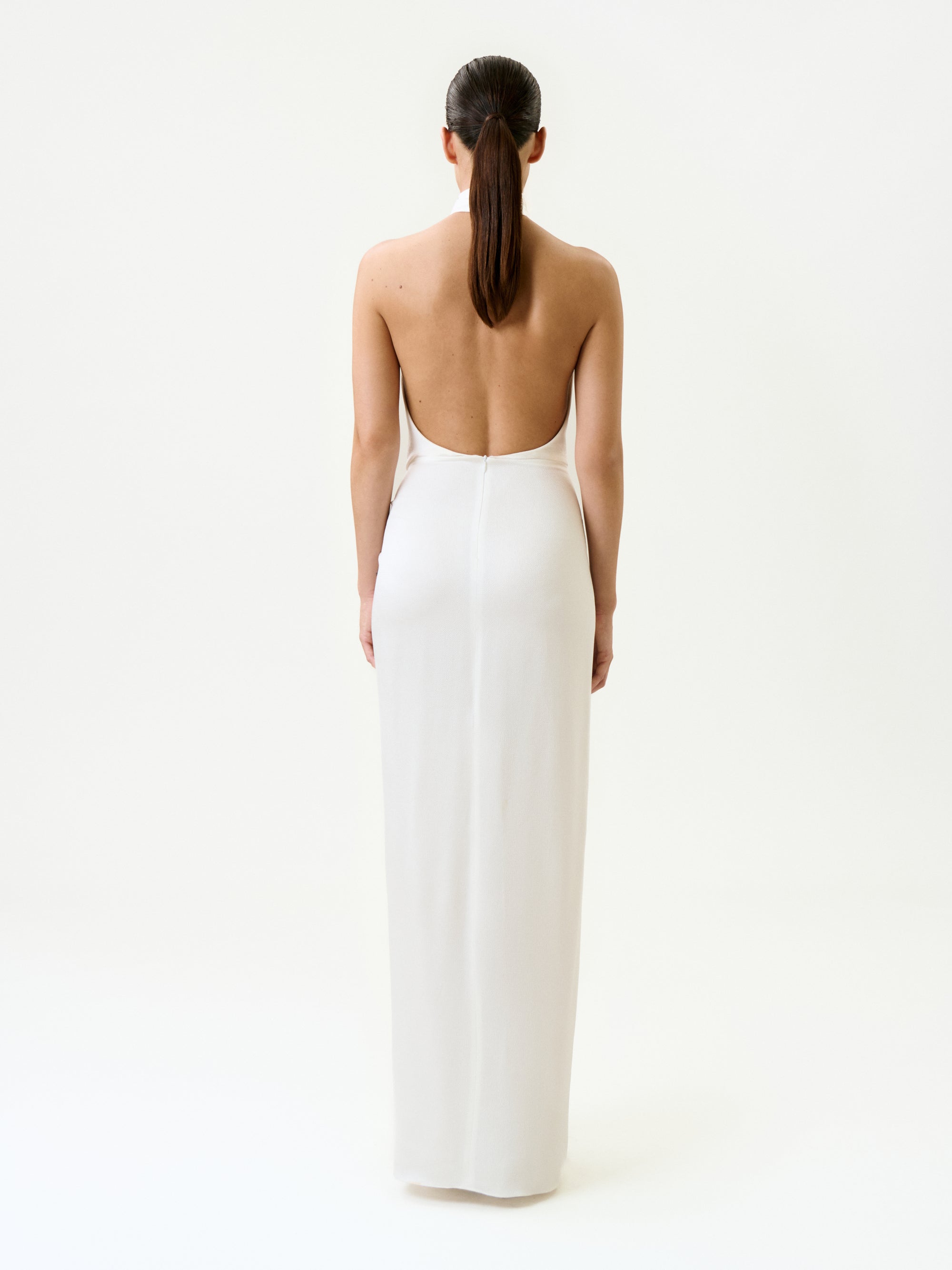 A woman with dark hair in a low ponytail stands with her back to the camera, wearing the Marisela Dress—a long, white maxi dress made in Portugal—showcased against a minimalist plain white background.