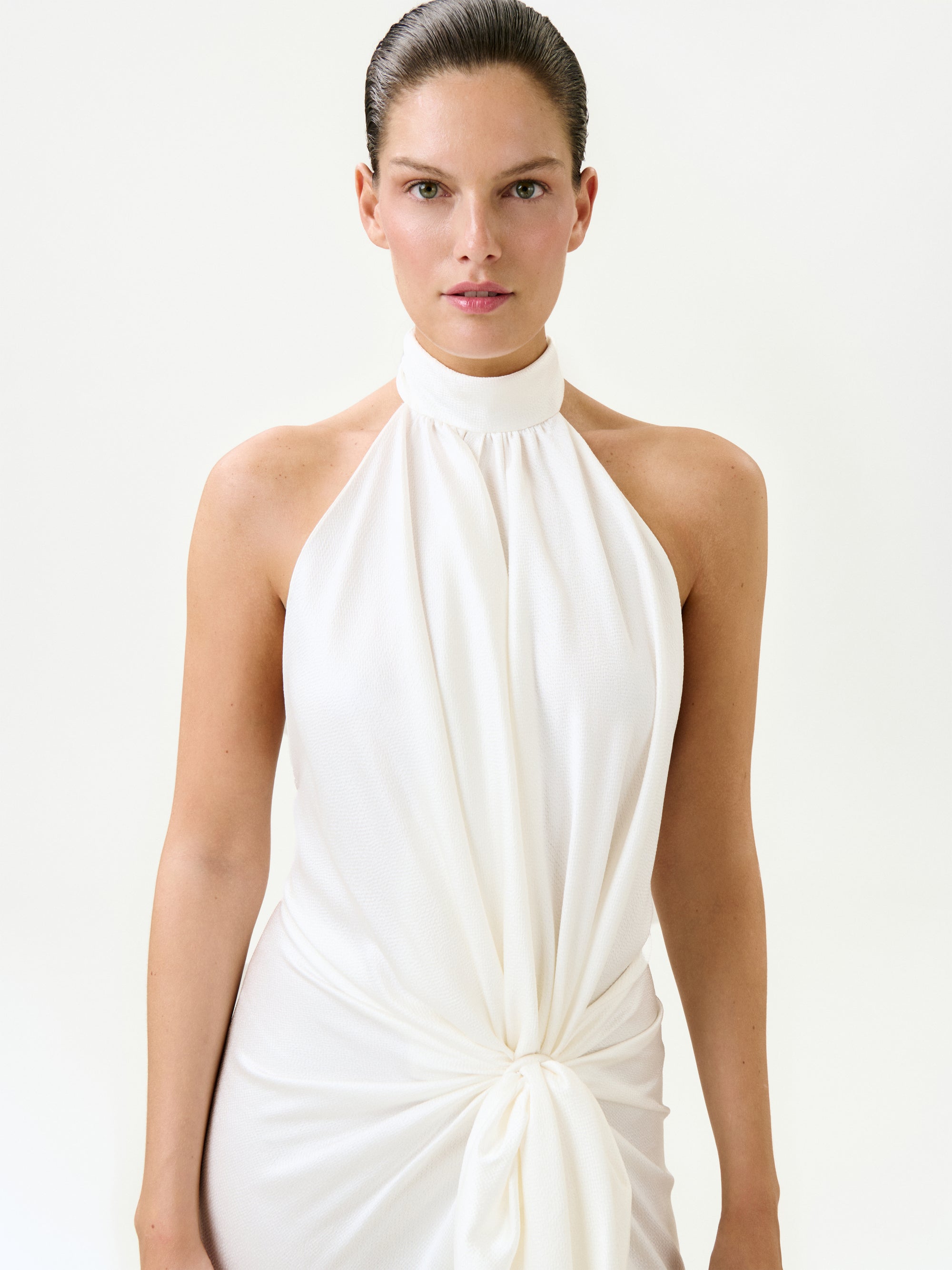 A woman with slicked-back hair models the Marisela Dress—a sleeveless white halter-neck maxi made in Portugal with a knotted waist detail—standing against a plain light background.