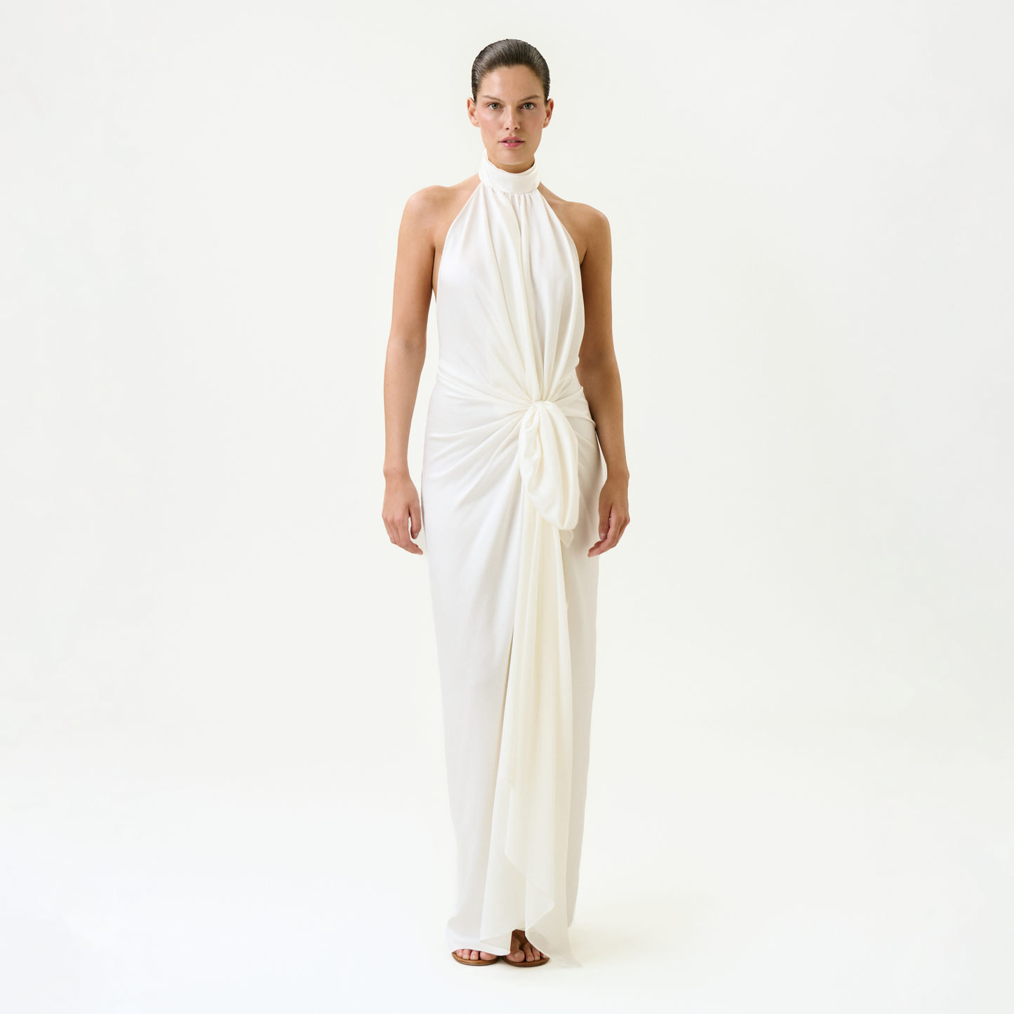 A woman stands facing forward in the Marisela Dress—a sleeveless, floor-length white maxi with a high neckline and draped knot waist, expertly crafted from fine Italian fabric—against a plain white background.