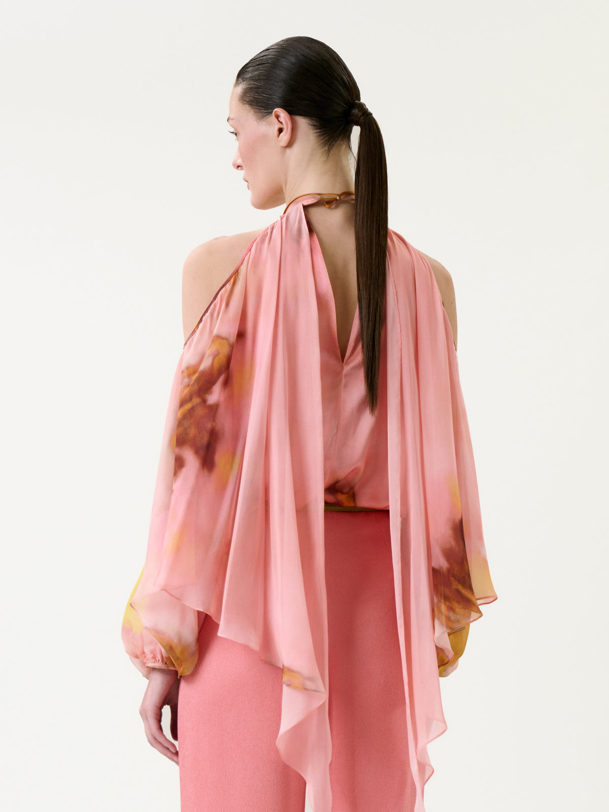 A woman with a sleek ponytail is turned away, highlighting the back details of the Shaan Blouse—a flowing, pink, off-the-shoulder top with long draped sleeves—paired with a matching skirt against a plain background.