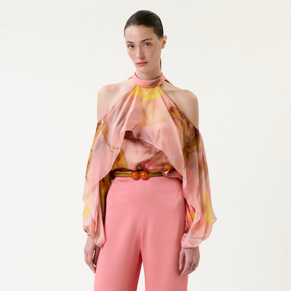 A woman wearing the Shaan Blouse—a colorful, flowing cold-shoulder top—paired with pink pants stands against a plain white background, looking at the camera with a neutral expression.