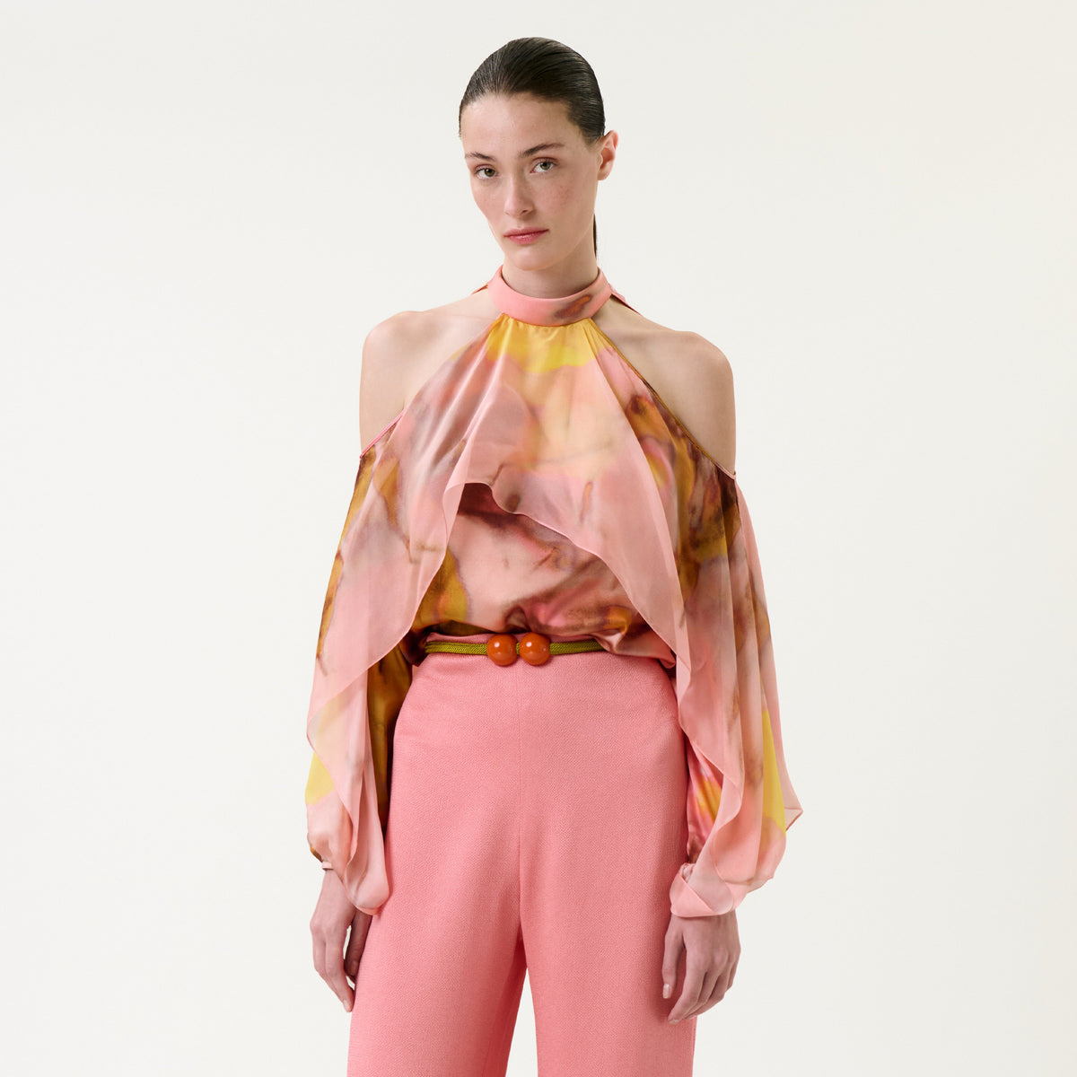 A woman wearing the Shaan Blouse—a colorful, flowing cold-shoulder top—paired with pink pants stands against a plain white background, looking at the camera with a neutral expression.