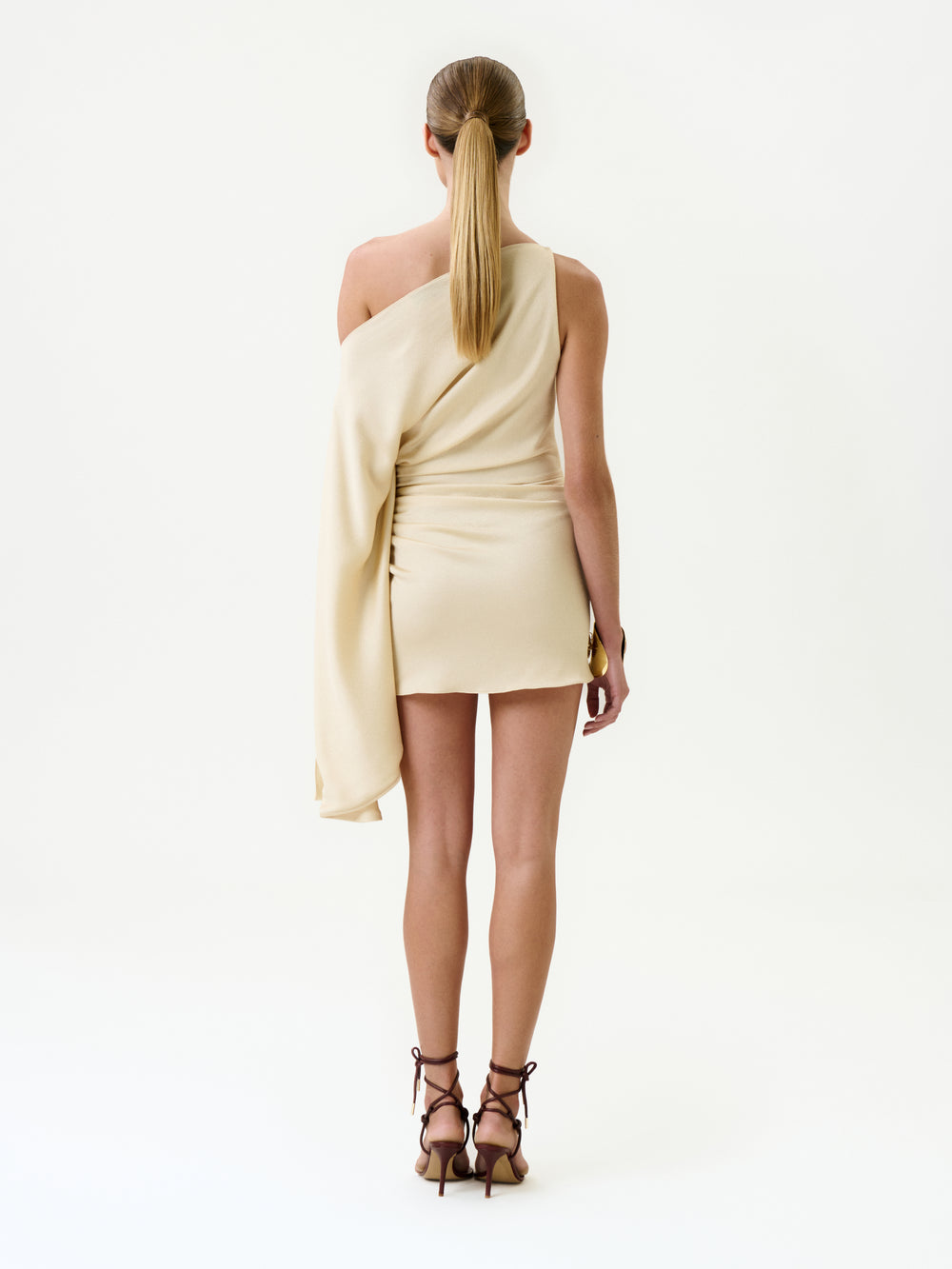 A woman faces away in the Nia Dress, a light beige one-shoulder mini crafted from luxurious Italian fabric with an asymmetrical drape. Her hair is in a sleek ponytail and she wears brown high-heeled strappy sandals against a plain white background.