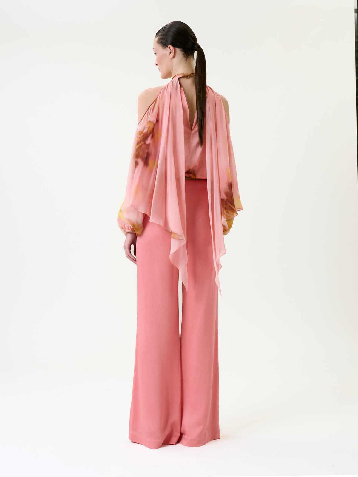 A woman stands facing away, wearing the Palermo Pant in pink with wide legs and a flowy, floral blouse featuring draped sleeves. Her hair is styled in a sleek ponytail against a plain white background.
