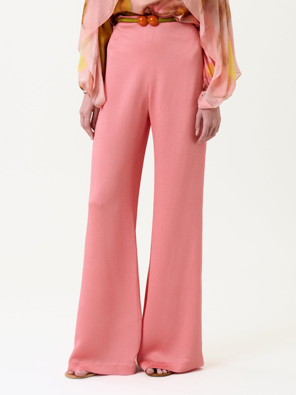 A person wearing the Palermo Pant—high-waisted, wide-leg pink pants styled with a brown belt and a vibrant, flowy blouse. Only the lower half of the body is visible.