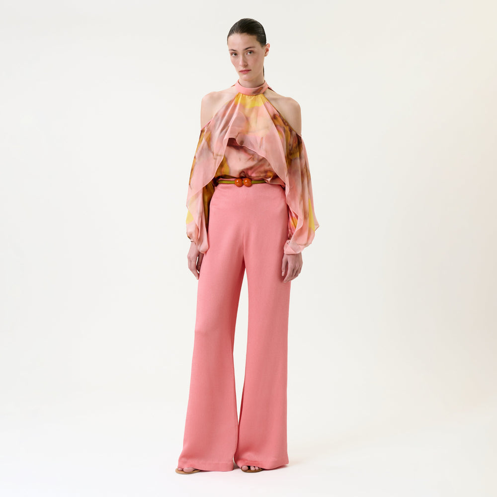 A woman stands against a white background wearing the Palermo Pant in pink and a flowy, multicolor halter top with open shoulders. She has slicked-back hair and a neutral expression.